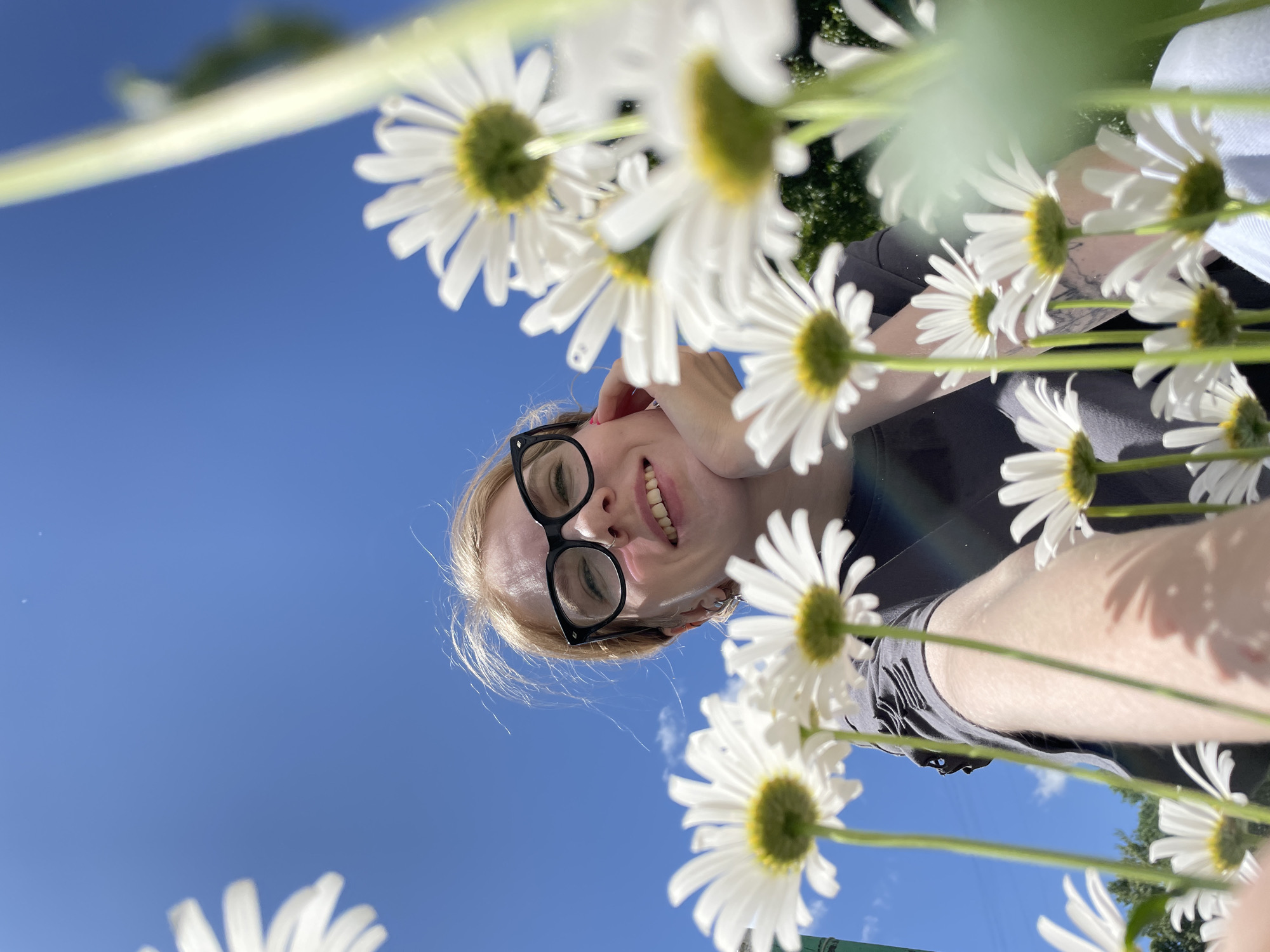 A low-angle outdoor selfie features a young woman with blonde hair and glasses, smiling and resting her chin on her hand. She is surrounded by several white daisies in the foreground, with a bright blue sky and some trees in the background. The woman is wearing a dark grey t-shirt. The overall impression is of a sunny day. The specific location within Izhevsk, Russia cannot be determined from the image alone.