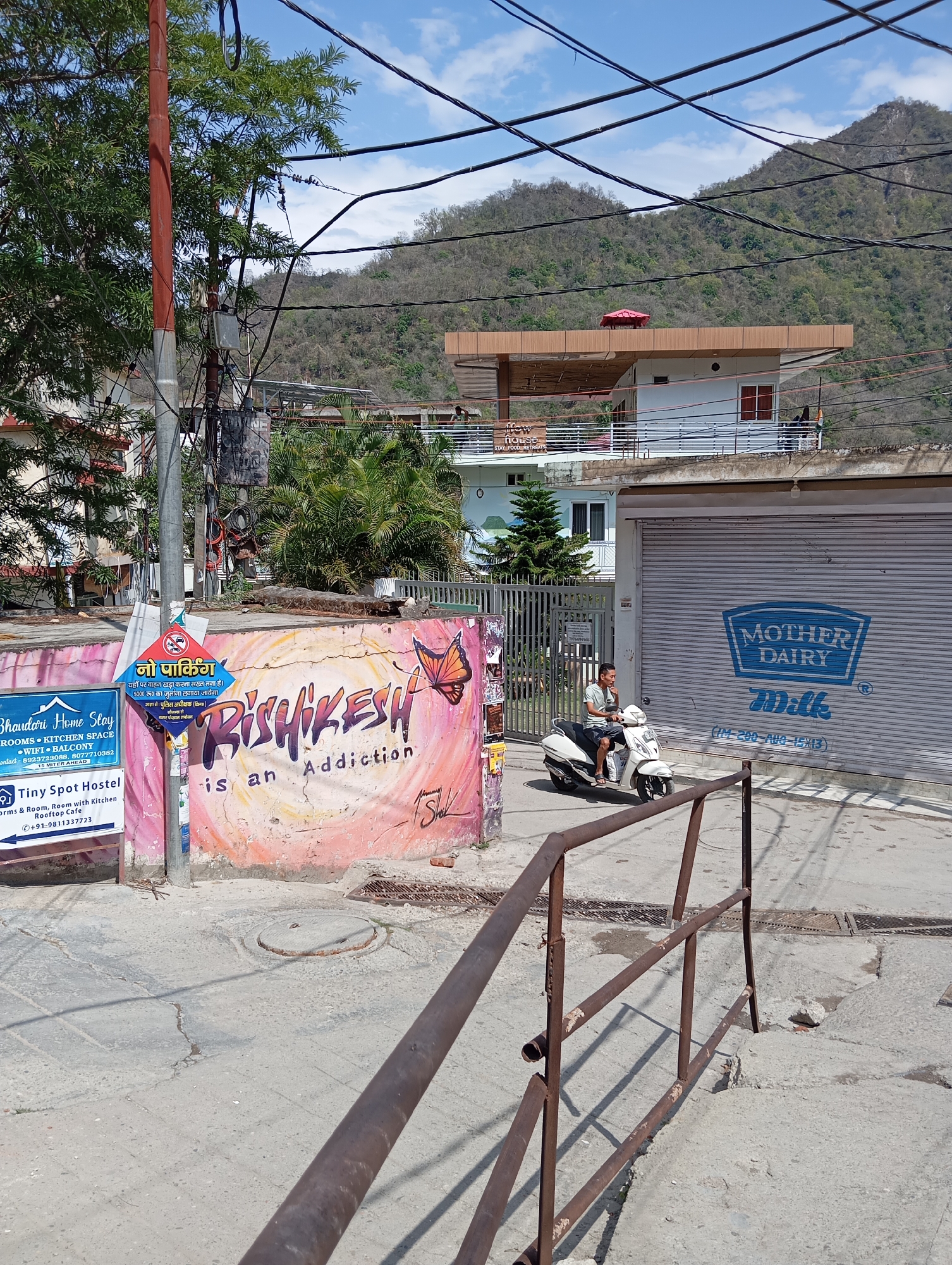 The image shows a street scene in Rishikesh, India. A man is riding a white scooter past a storefront with a "Mother Dairy" logo on the closed shutter. To the left, a painted wall displays the words "Rishikesh is an Addiction" along with advertisements for "Bhandari Home Stay" and "Tiny Spot Hostel." Above this wall is a "No Parking" sign in Hindi. A rusty metal railing is visible in the foreground, while mountains rise in the background under a partly cloudy sky. Overhead, numerous electrical wires crisscross the scene.
