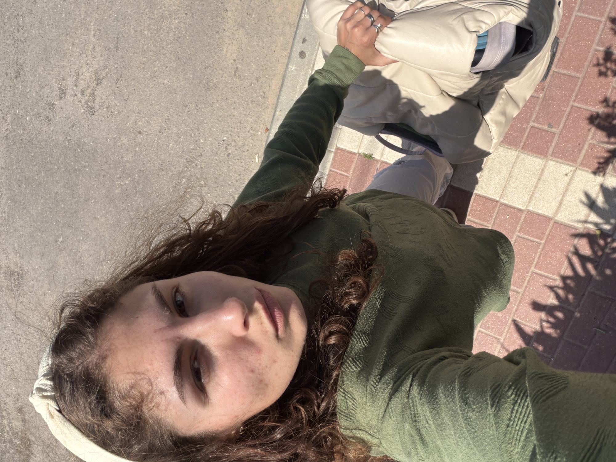 A first-person, high-angle photograph shows a person on an outdoor sidewalk in Kiryat Bialik, Israel. The person is wearing a textured, long-sleeved, olive green shirt. The person is also wearing a white headband. They have curly brown hair. The person's face is visible, displaying freckles and minor skin imperfections. Their left hand holds a beige, quilted bag. The ground consists of gray concrete on the left and reddish-brown brick tiles on the right. The shadow of a tree is cast on the brick tiles.