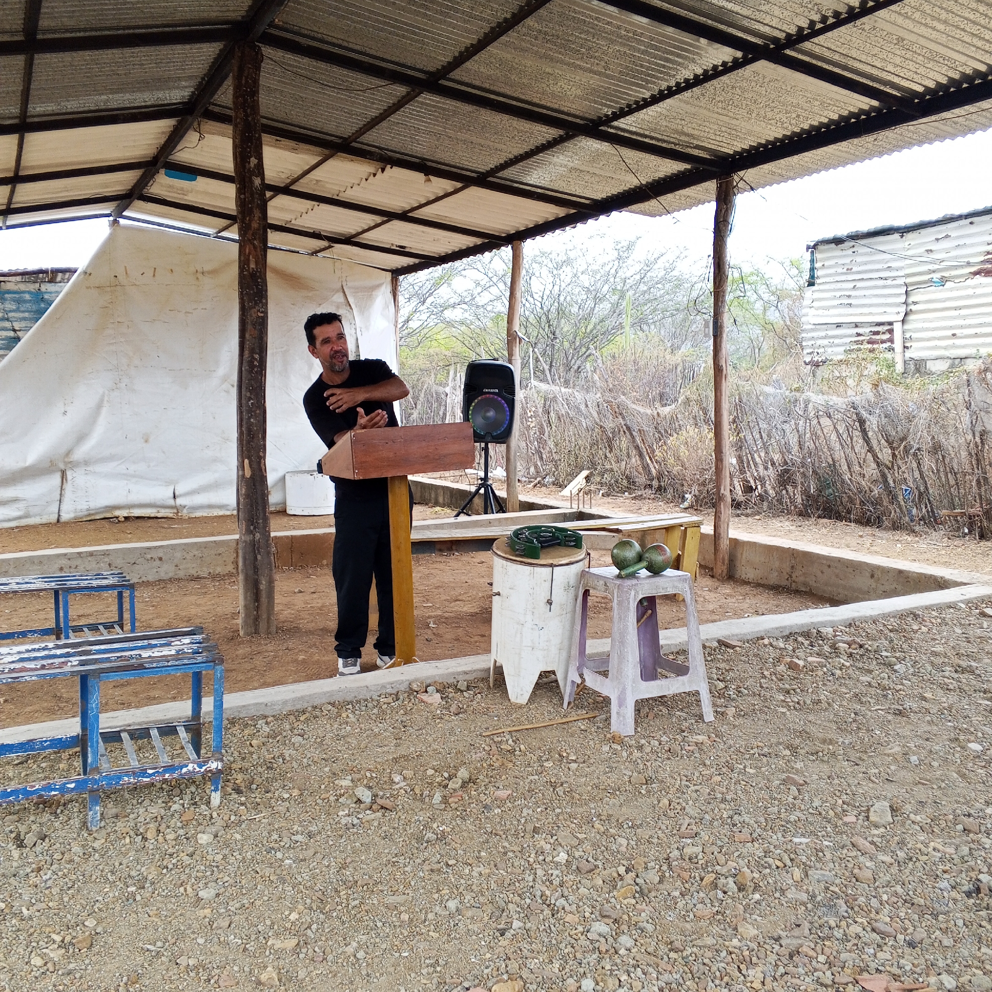The image shows a man standing behind a wooden lectern under a makeshift open-air structure in an unidentified location. The structure has a corrugated metal roof supported by wooden posts, providing partial shade. The man is wearing a black shirt and pants with white sneakers, and he is gesturing with his hands as if speaking or presenting. Behind him is a portable speaker on a tripod stand. To his right, a white cylindrical container is visible with a percussion instrument on top, next to a small stool with two avocado-shaped objects on it. In the foreground to the left, there are some blue benches. The ground is covered in gravel, and there is a low concrete border outlining the space. The background reveals a rural or semi-rural environment with sparse vegetation and a fence line made of branches and wires. A dilapidated building is visible in the upper right corner of the frame.
