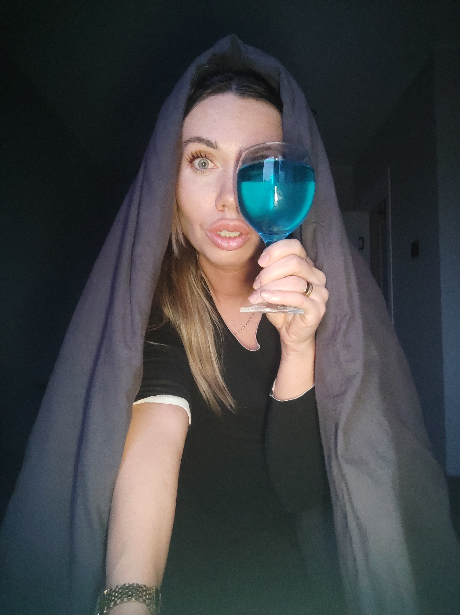 A woman poses for a selfie while holding a glass of blue liquid in front of her left eye. She is draped with a gray sheet. She has blond hair and wears a black shirt. A gold bracelet is visible on her left wrist. She has a ring on her ring finger. The location is indoors, possibly a bedroom. Based on the presence of a doorframe in the background, the location is residential and does not appear to be in the city of Votkinsk, Russia.
