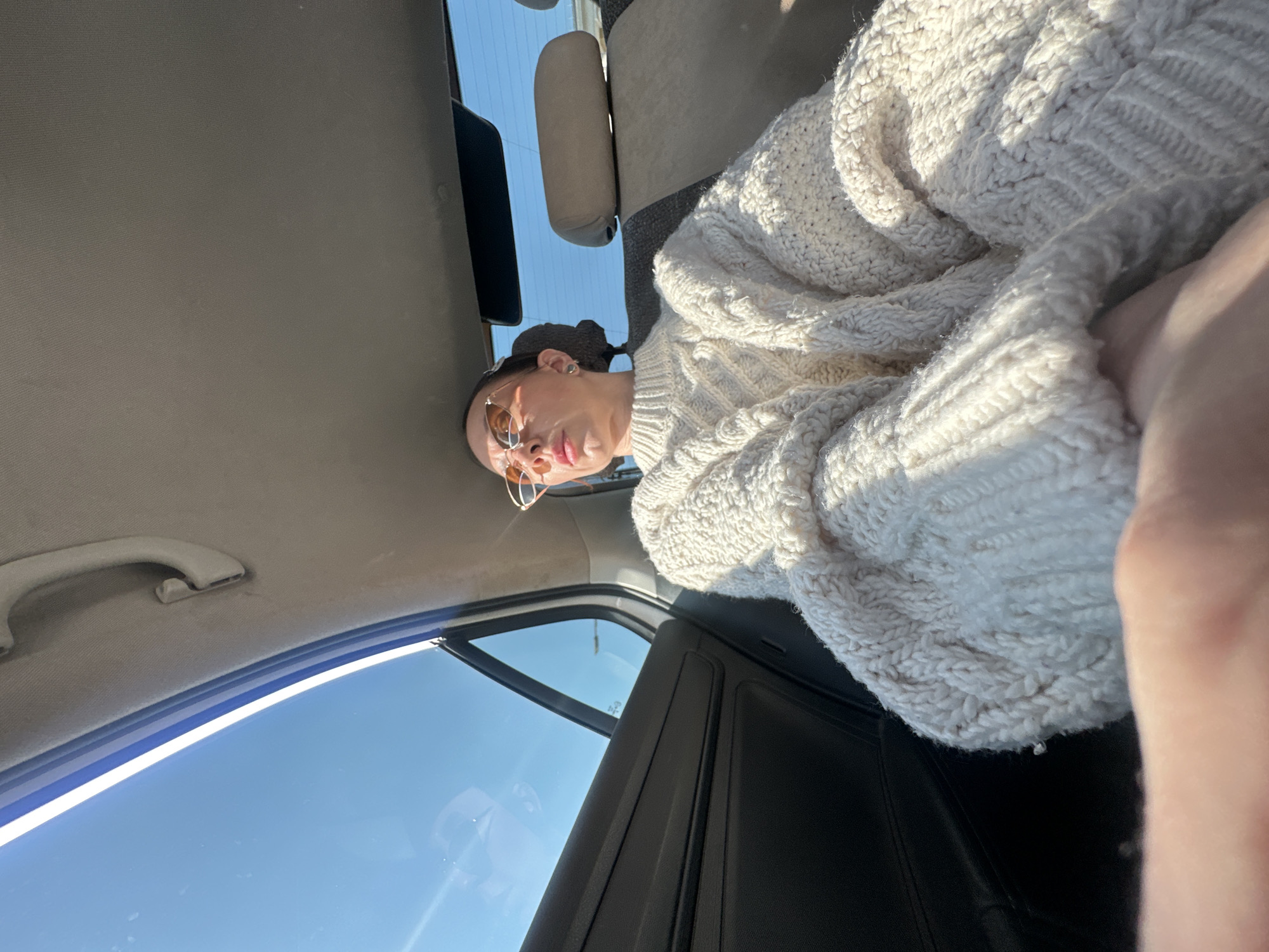 The image shows a person inside a car. The person is wearing sunglasses and a white cable-knit sweater. The car's interior is visible, including the headrest, window, and roof handle. The window shows a clear blue sky. The location is unknown and there is no information to suggest the city of Votkinsk, Russia is the location.