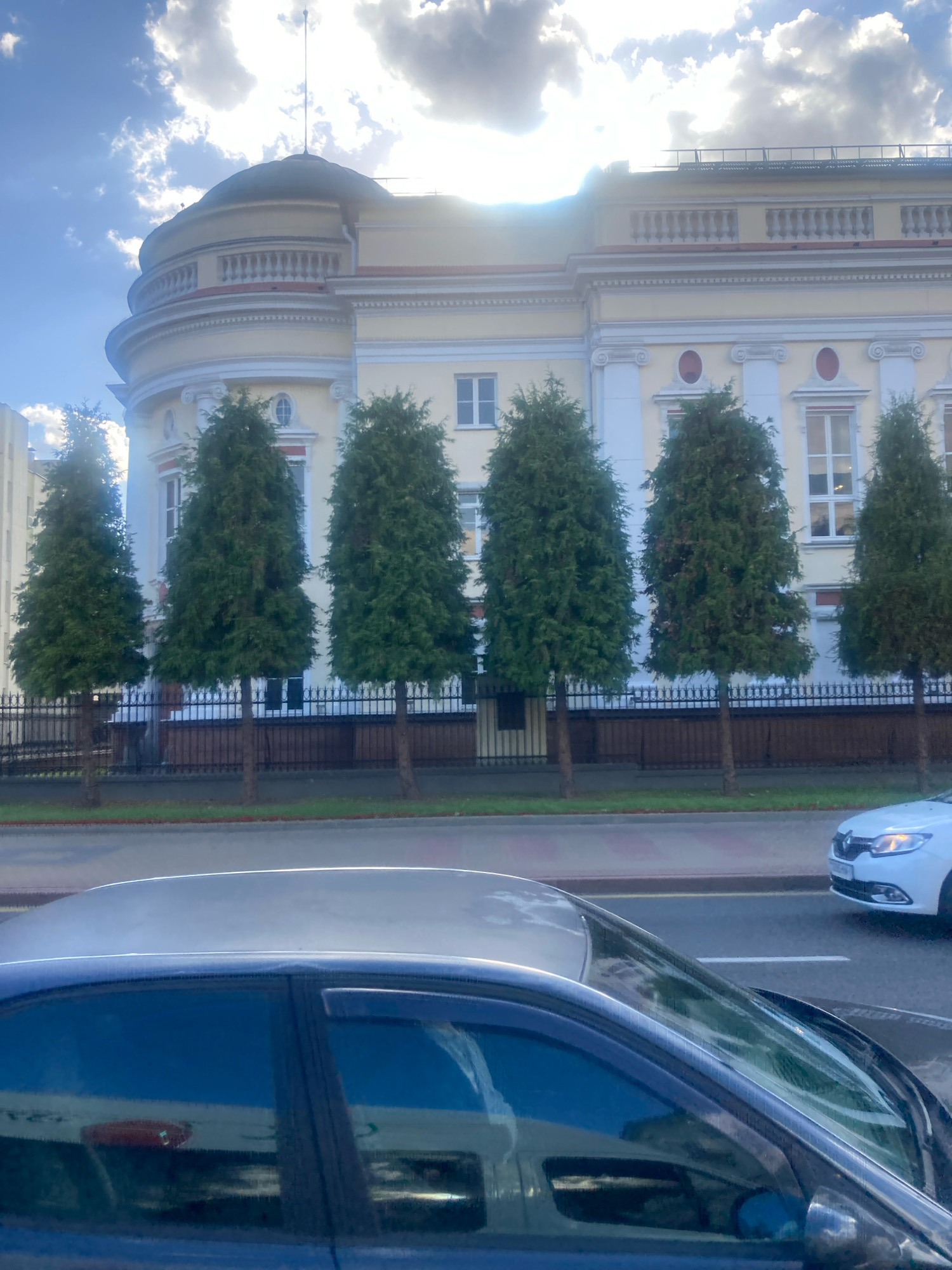 The image shows a multi-story building with a rounded section topped by a dome, and a row of manicured evergreen trees in front of it. The building appears to be a public institution, possibly an educational or governmental building, given its formal architecture with columns and decorative elements. The sky is cloudy with patches of blue, and sunlight is filtering through, creating a bright atmosphere. In the foreground, there are cars on a paved road, suggesting an urban setting. The scene captures a bright, slightly overcast day in a city. The building's style and the surrounding environment are consistent with architecture found in Brest, Belarus.