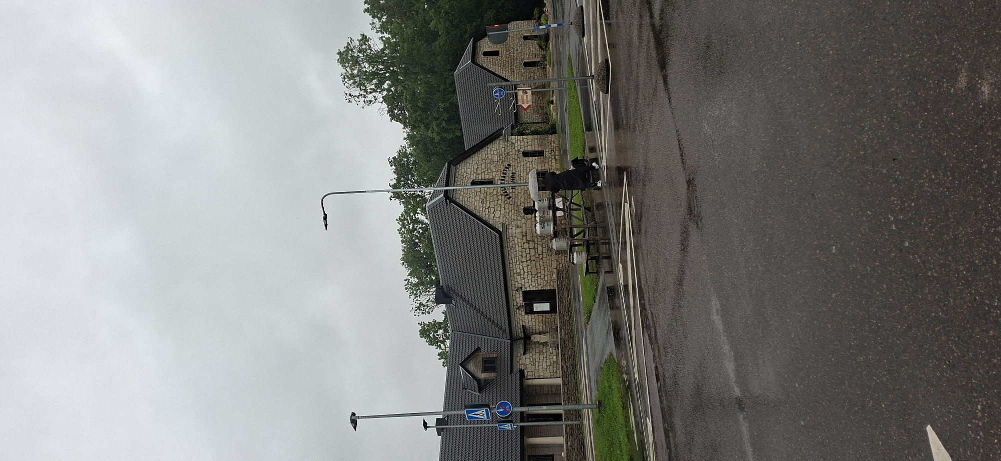 The image depicts an outdoor scene on a cloudy day, likely in Kaali, Estonia. In the foreground, a wet asphalt road with white lane markings is visible. A pedestrian crossing is marked on the road. To the left of the road, there is a grassy area with a stone wall. Beyond the wall, a stone building with a dark, pitched roof is situated. The building has several windows and at least two doorways.
To the left of the building, a lamppost stands near a pedestrian crossing sign. In front of the building, there is a wooden picnic table laden with several large, metallic milk churns. A person wearing a dark coat and holding a clear umbrella stands near the table, seemingly interacting with the churns. Another lamppost is visible to the right of the building. In the background, lush green trees form a dense line. Further to the right, another similar stone building is partially visible, and a red and white sign on a post is present. The overall impression is of a rural or semi-rural setting, possibly a farm or a visitor center, under overcast conditions.