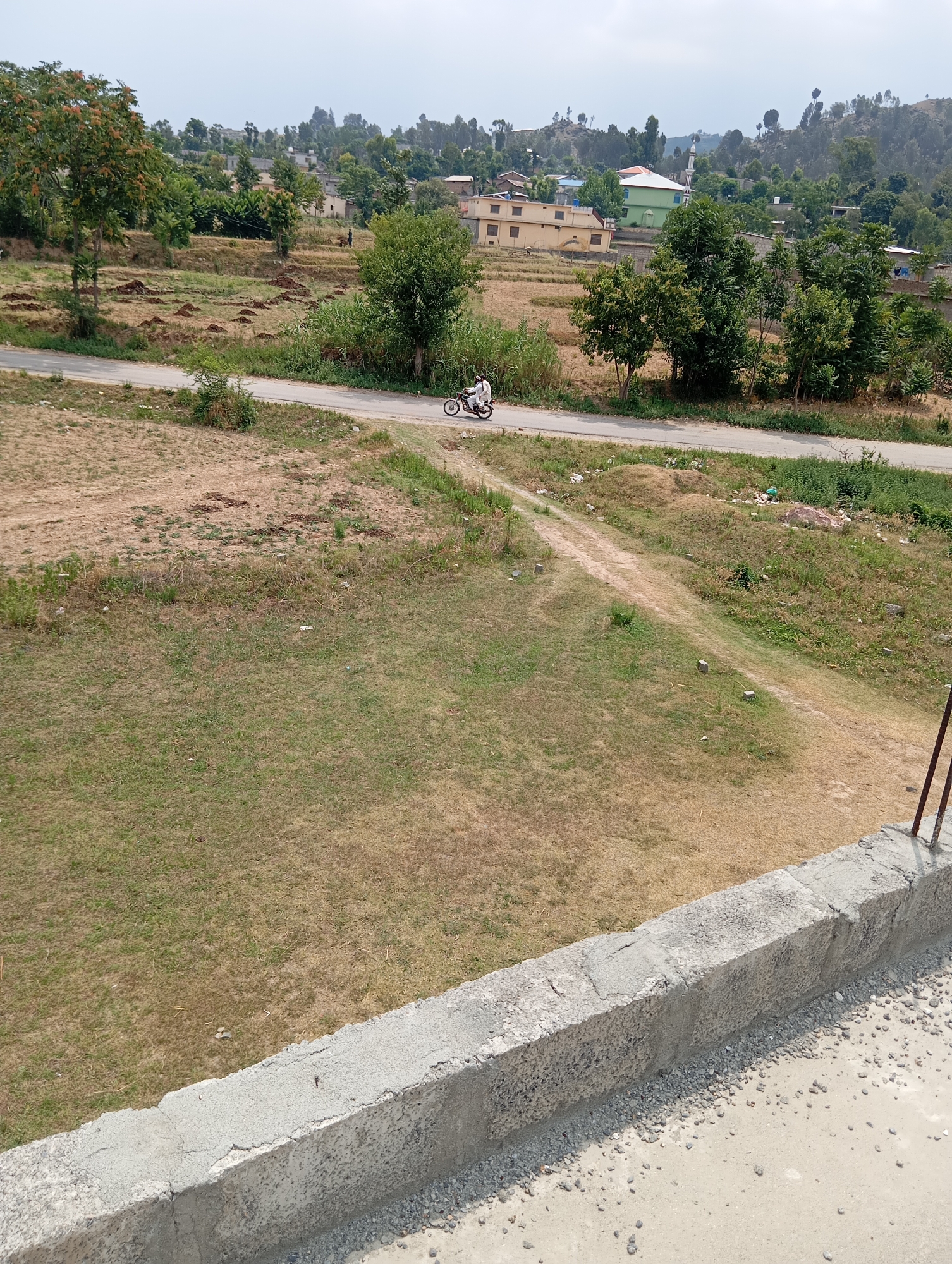 A high-angle, eye-level shot captures a rural scene on an overcast day. In the foreground, a concrete wall, appearing to be part of a building's foundation or parapet, runs horizontally across the bottom of the frame. The wall is rough and textured, with small pebbles embedded in its surface and a lighter concrete base. Beyond the wall, a gently sloping grassy field dominates the midground. The grass is a mix of green and dry, yellowing patches, with some scattered debris, including small stones and what appears to be dried leaves. A dirt path winds through the grass, leading towards a paved road in the distance. On the road, a person on a motorcycle is traveling to the left. The road is lined with dry, sparse vegetation on one side and a more densely vegetated embankment on the other. In the background, a cluster of buildings, including houses and a mosque with a green roof and a minaret, are nestled among trees on a hillside. The landscape is characterized by rolling hills covered in trees and vegetation, creating a sense of a remote, undeveloped area. The sky is a pale, hazy grey, indicating a cloudy or foggy atmosphere.