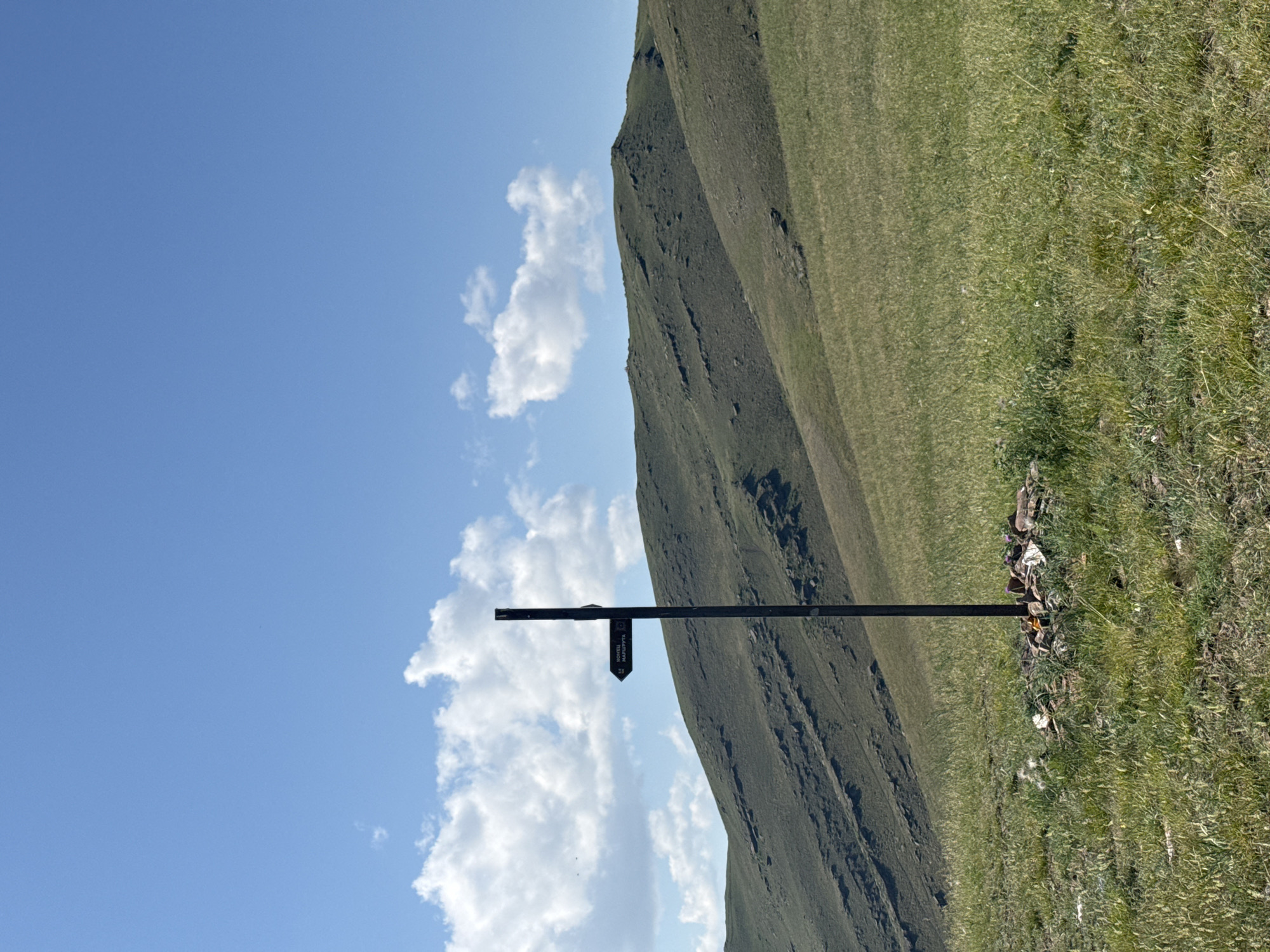 An outdoor, eye-level, full shot captures a remote, grassy hillside under a bright blue sky dotted with fluffy white clouds. In the foreground, a solitary, dark, metal signpost stands planted in a circle of small, rough stones. A single arrow-shaped sign is attached to the post, pointing left, with dark text on a lighter background, possibly indicating a trail or direction. The sign has a small symbol that looks like a circular path with a dot in the center. Behind the signpost, the landscape transitions into rolling, green hills that recede into the distance. The grass in the foreground is a mix of green and yellow, with some taller stalks, and small clumps of wildflowers are visible near the base of the signpost. The image depicts a natural, undeveloped environment, suggesting a rural or mountainous area. Based on the provided information, the location is identified as the city of Ust'-Abakan, Russia.