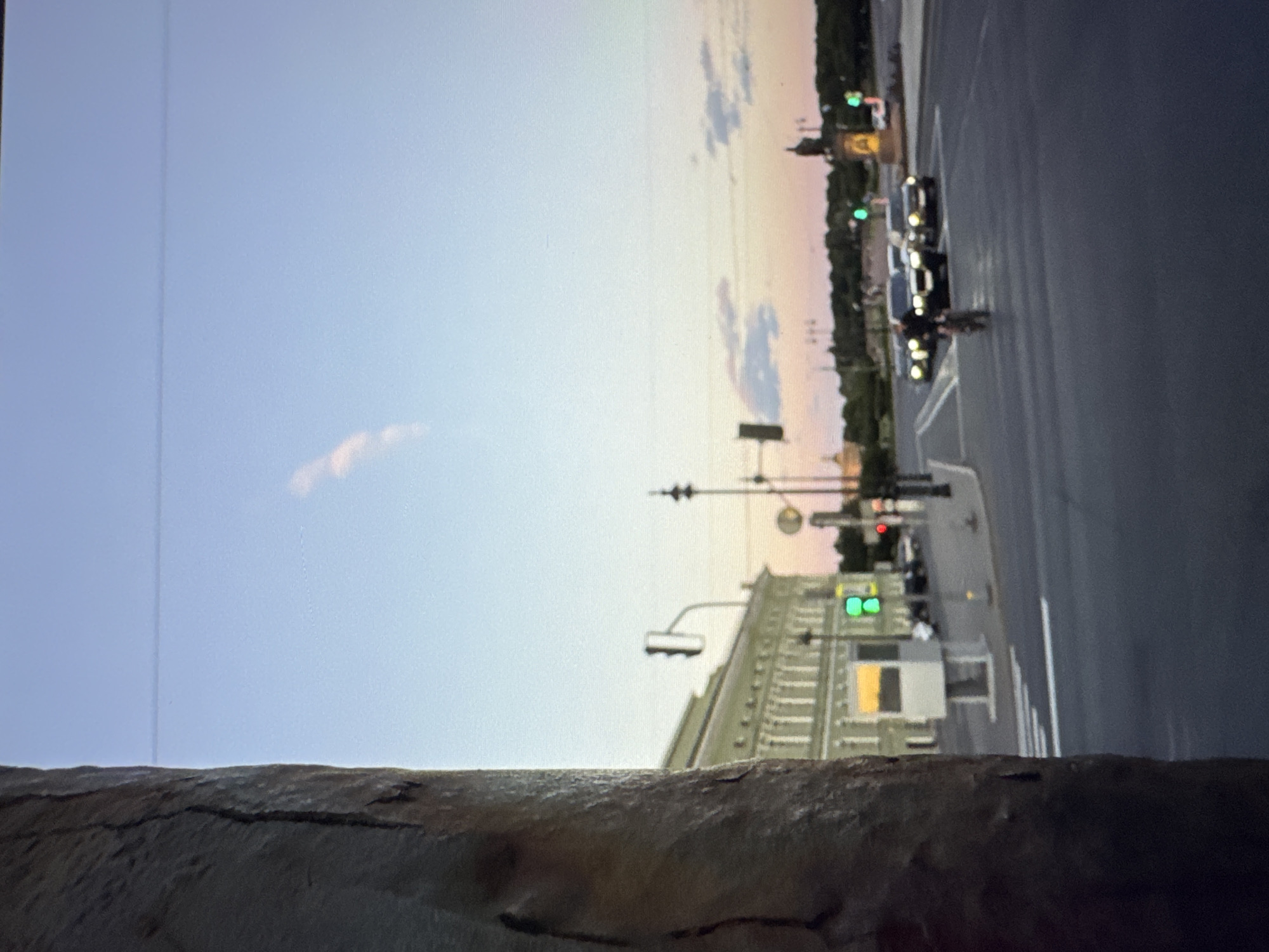 The image shows a street scene in a city at dusk. In the foreground on the left, a textured, dark, vertical surface, possibly a pillar or wall, occupies a significant portion of the frame. The background is a city street with a large, light-colored building on the left side. This building has multiple windows and architectural details. A traffic light with a green signal is visible. Across the street, several cars are driving, and a person is riding a bicycle. In the distance, trees line the horizon, and a statue or monument can be seen. The sky is a gradient of light blue and soft orange, indicating either sunrise or sunset. There are a few wispy clouds in the sky. The overall scene is captured from a slightly elevated perspective, looking across the street and towards the buildings and distant landscape.