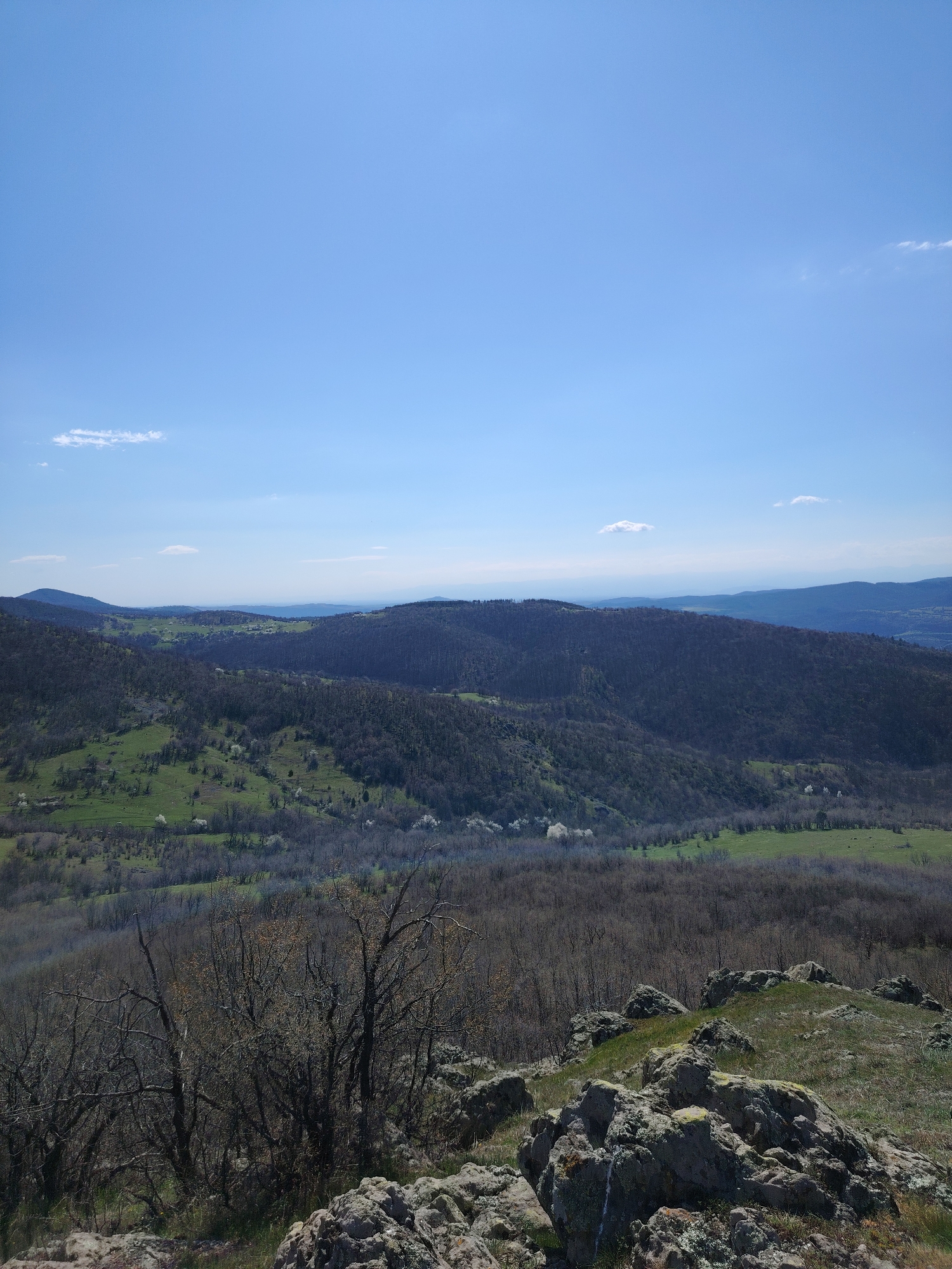 The image shows a landscape in Bata, Bulgaria. In the foreground are rocks and sparse vegetation on a hilltop. Behind, a valley stretches between rolling hills covered with trees and patches of green meadow. The sky is blue with a few scattered clouds. The environment is natural and appears to be a rural or mountainous area. There are no people visible in the image.