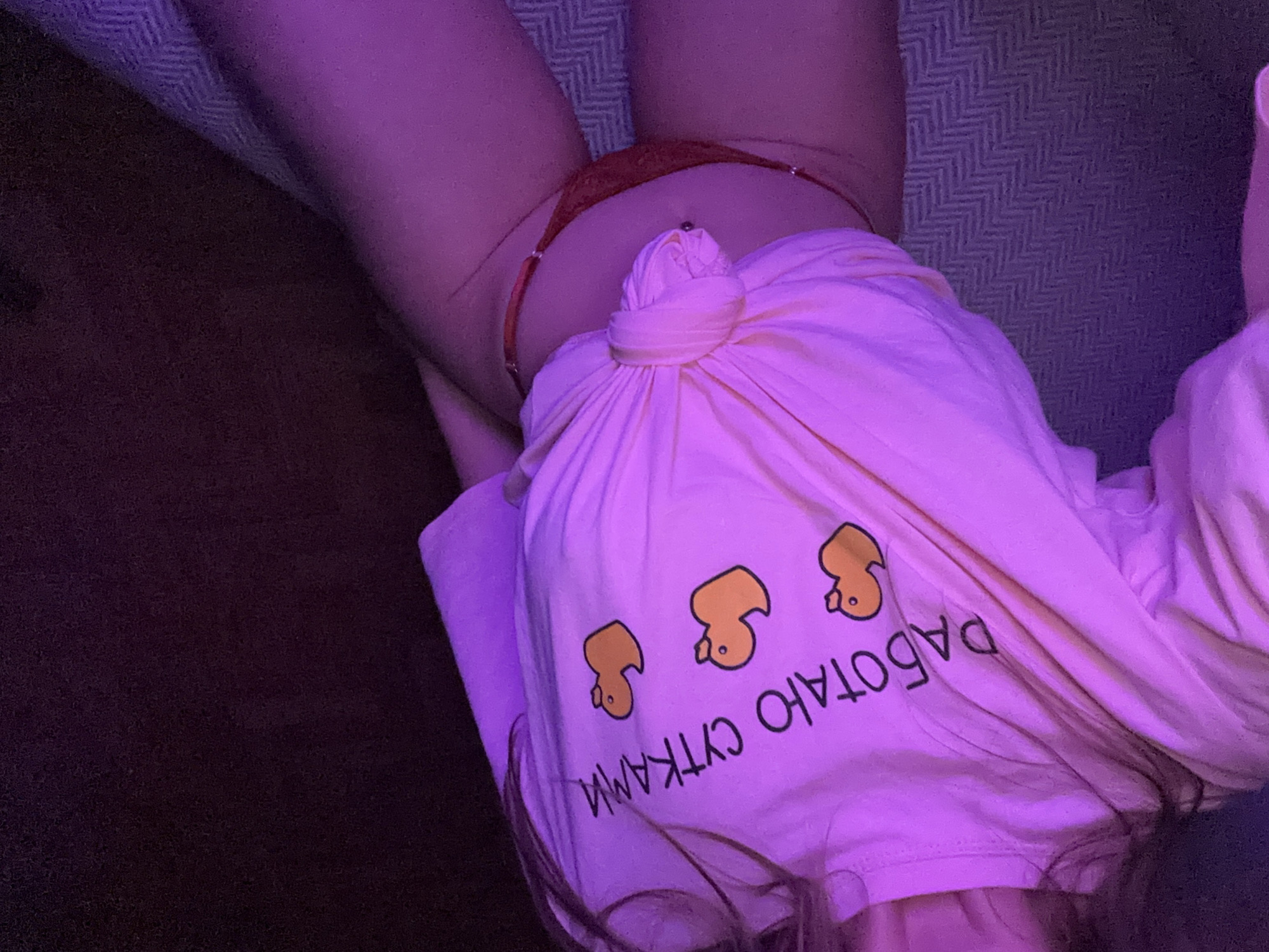 The image shows a person wearing a pink t-shirt with a knot tied at the waist. The t-shirt has a print of three yellow rubber ducks and text in Russian which reads "РАБОТАЮ СУТКАМИ" (I work around the clock). The person is also wearing red underwear. The lighting in the image is purple, suggesting it might be nighttime or a room with colored lighting. The background is dark and appears to be a textured surface, possibly a bed or couch. There is no visible indication of the location being in the city of Khimki, Russia.