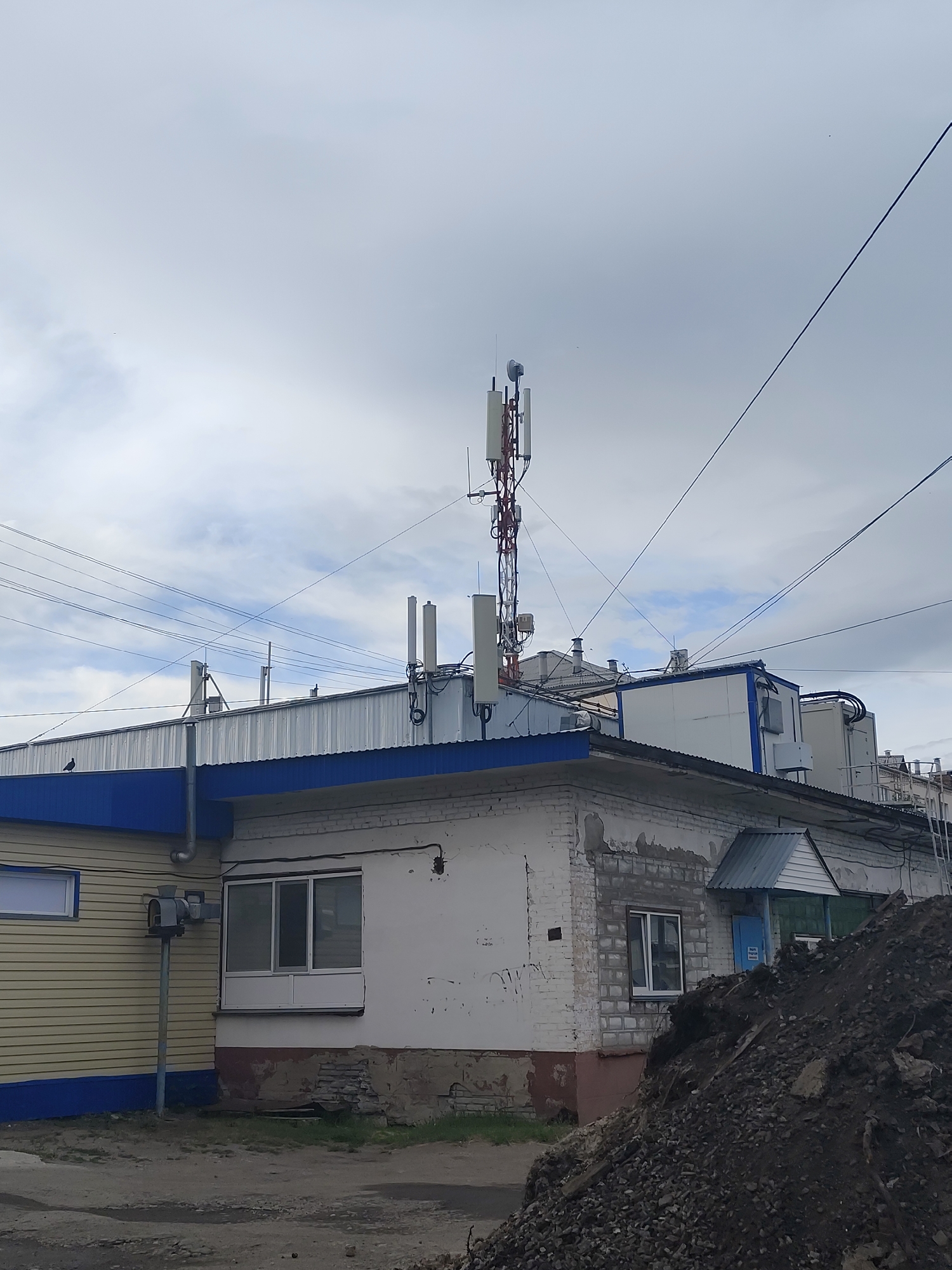 The image shows a cell tower against a cloudy sky. The tower is tall and red and white, with antennas on top. In the foreground, there is a brick building with a blue and yellow facade. A pile of dirt and rocks is in the lower right corner of the image. The scene appears to be in an industrial or commercial area, with power lines strung across the sky. The overall impression is one of urban utility infrastructure.