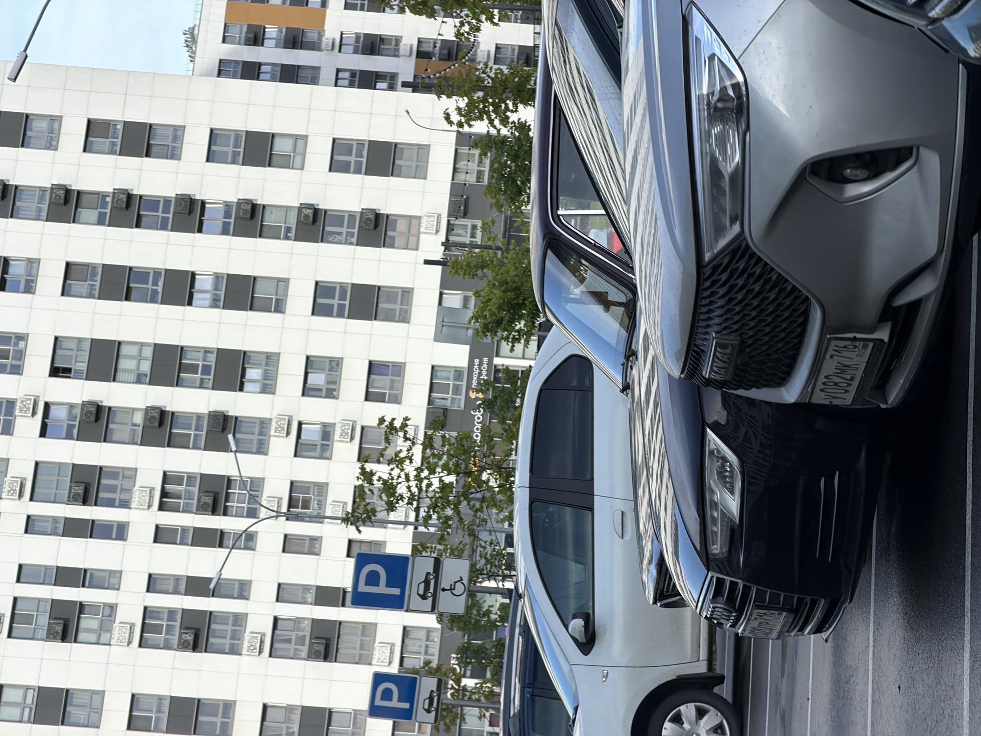 In Kazan, Russia, a frontal, eye-level shot captures a parking lot in front of a large, modern residential building. The building, characterized by its white facade and numerous windows, dominates the background. Three cars are visible in the foreground, partially obscuring the lower floors of the building and street signs. A white car is parked on the left, a dark sedan is in the middle, and a light-colored SUV is on the right. Two blue parking signs are mounted on a pole to the left of the building. One sign displays a white "P" for parking, and a smaller sign below it shows a silhouette of a car. Another sign nearby indicates parking for disabled individuals with a wheelchair symbol. The sky above is overcast and a pale blue.