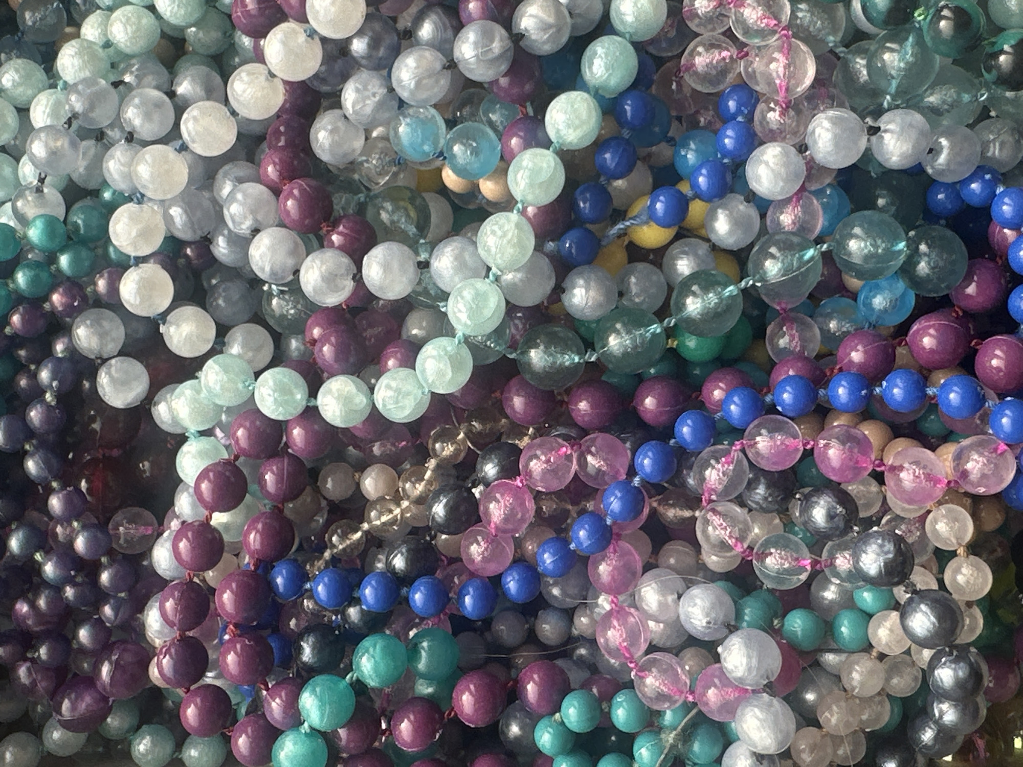 The image is a close-up, overhead shot of a large pile of colorful, round beads strung on threads, creating multiple strands. The beads vary in size, color, and opacity. Colors include shades of purple, blue, green, white, and pink, with some beads being translucent and others opaque. The lighting appears to be diffused, creating soft shadows. There are no people or animals visible in the image. The background is completely filled with the beads, suggesting they are in a container or spread out on a surface. The location is unknown.