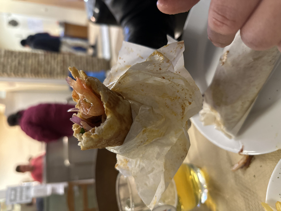 This image captures a close-up, somewhat askew view of a partially eaten wrap or sandwich in a casual dining setting in Nea Erithrea, Greece. The main subject is the wrap, held by a hand in the foreground. It appears to be a flatbread or pita filled with fresh ingredients, notably red slices of tomato and thin, purple slivers of red onion. The wrap is loosely encased in white paper, which is stained with orange-brown residue, suggesting spices or sauce from the filling.
In the blurred background, several people are visible, indicating an active eatery. One individual in a dark blue top is further in the distance, while another person wearing a maroon or dark red jacket is seated closer to the mid-ground. A third person in a pink top can also be dimly seen. These figures suggest patrons enjoying meals or moving around in a restaurant, cafe, or similar establishment.
To the right of the wrap, a white plate with faint traces of food and a glass containing a bright yellow beverage, possibly juice, are partially visible. The lighting is consistent with an indoor setting, possibly artificial lighting during the day or evening. No text is visible in the image. The scene conveys a candid moment during a meal, with the focus sharply on the food in hand.