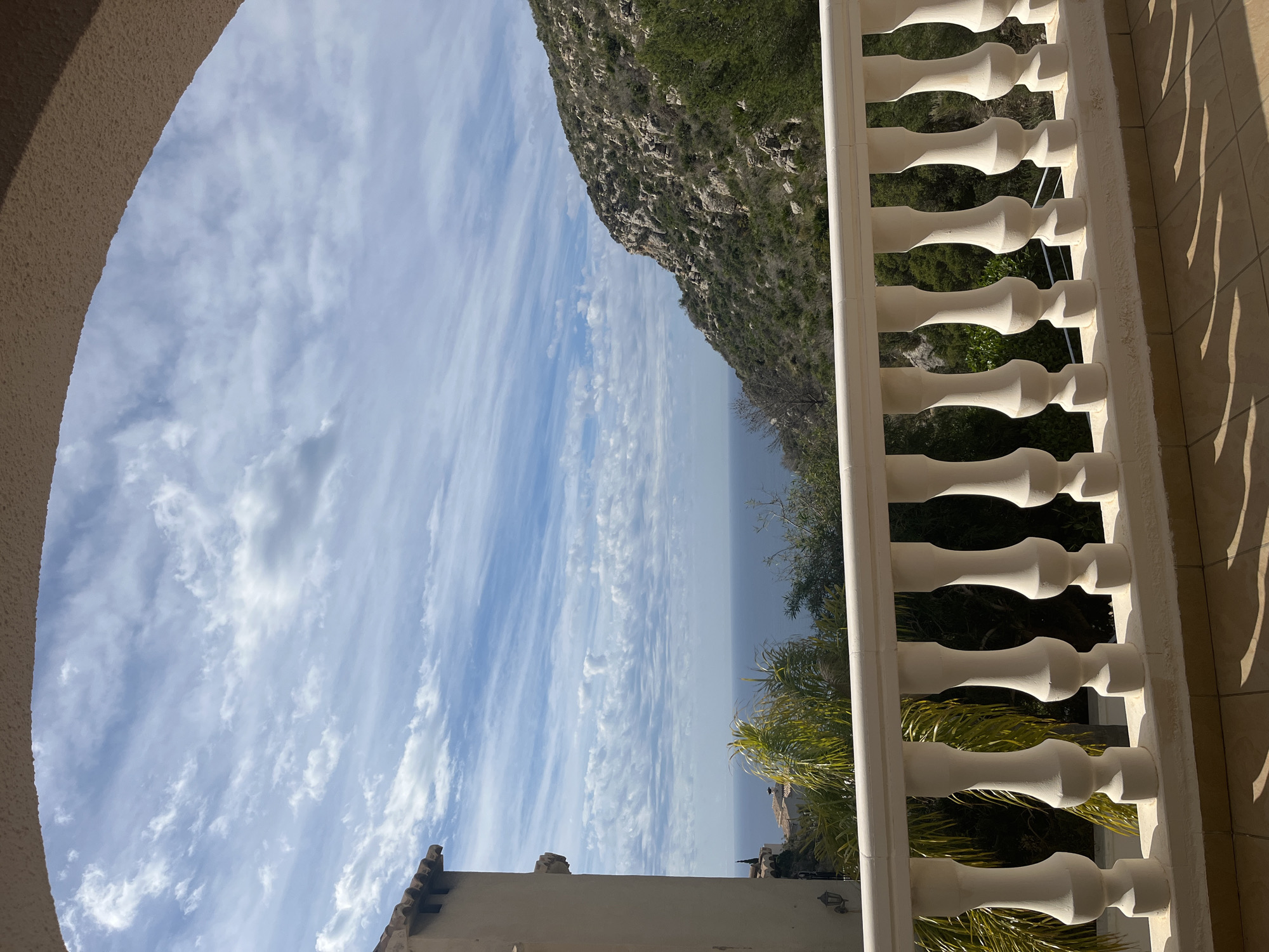 The image shows a view from what appears to be a balcony or terrace, framed by an arched structure and a white balustrade. The scene includes a stretch of the Mediterranean Sea meeting a rocky coastline covered with sparse vegetation. The sky is filled with light, streaky clouds. Part of a building with tile roofing is also visible in the lower part of the frame. The location is likely somewhere in el Poble Nou de Benitatxell / Benitachell, Spain, given the landscape.