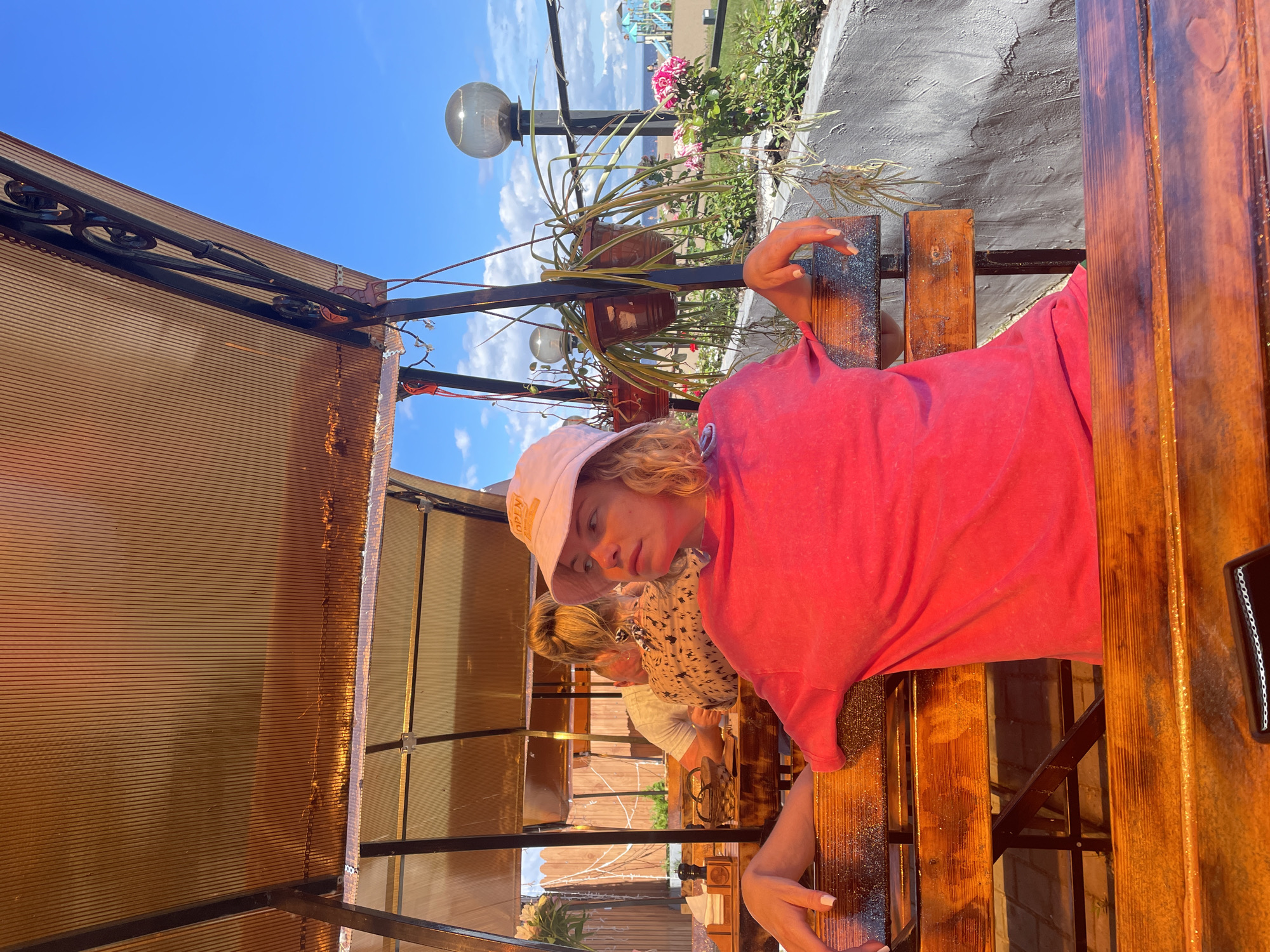 The image shows a young woman with blonde hair wearing a pink bucket hat and a bright pink t-shirt, sitting on a wooden bench under an orange translucent awning. She is looking to her right with a contemplative expression. Behind her, other people are visible at tables, and plants are hanging in pots. In the background, there is a gray concrete wall, a street lamp, and a view of the water and a playground. The scene appears to be an outdoor seating area of a cafe or restaurant. The image is likely taken in the city of Belozersk, Russia, given the context.