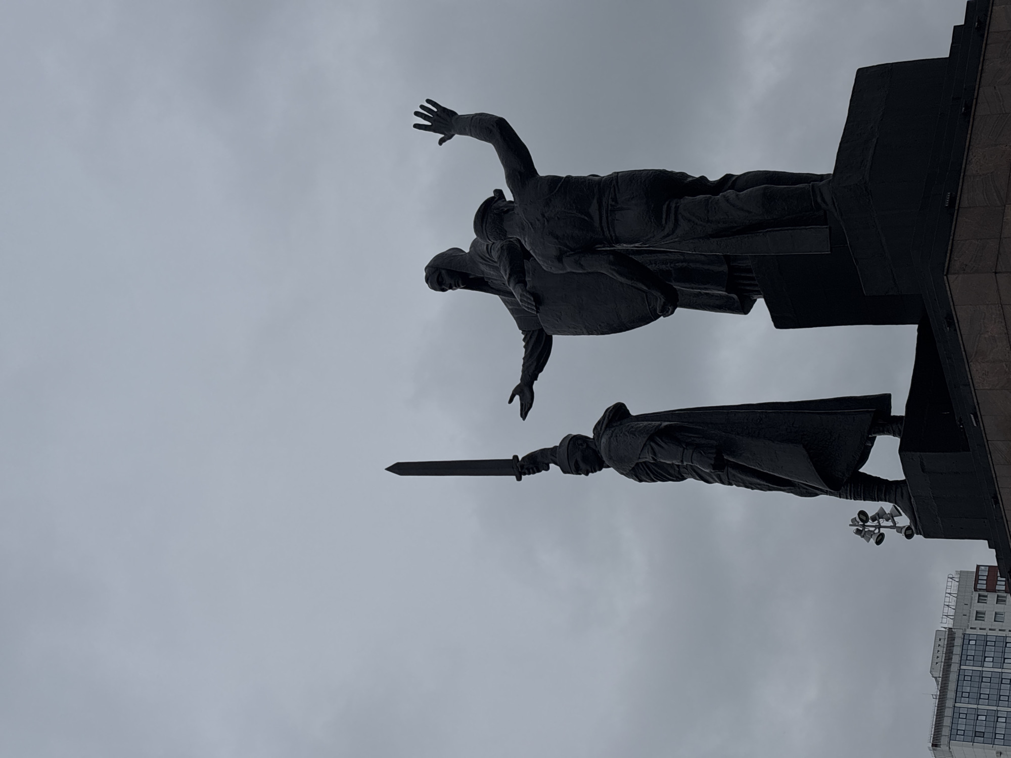 The image depicts a statue, possibly made of bronze or a dark-colored metal, against a cloudy sky. The statue appears to be a monument or memorial, possibly related to historical or military events. The statue features a male figure wielding a sword, standing beneath two figures locked in embrace. This is the Rear to the Front Monument in Perm, Russia. Part of a building, possibly an apartment or office complex, is visible in the lower right corner of the image. Street lights are visible near the statue. The overall lighting is subdued due to the overcast conditions.