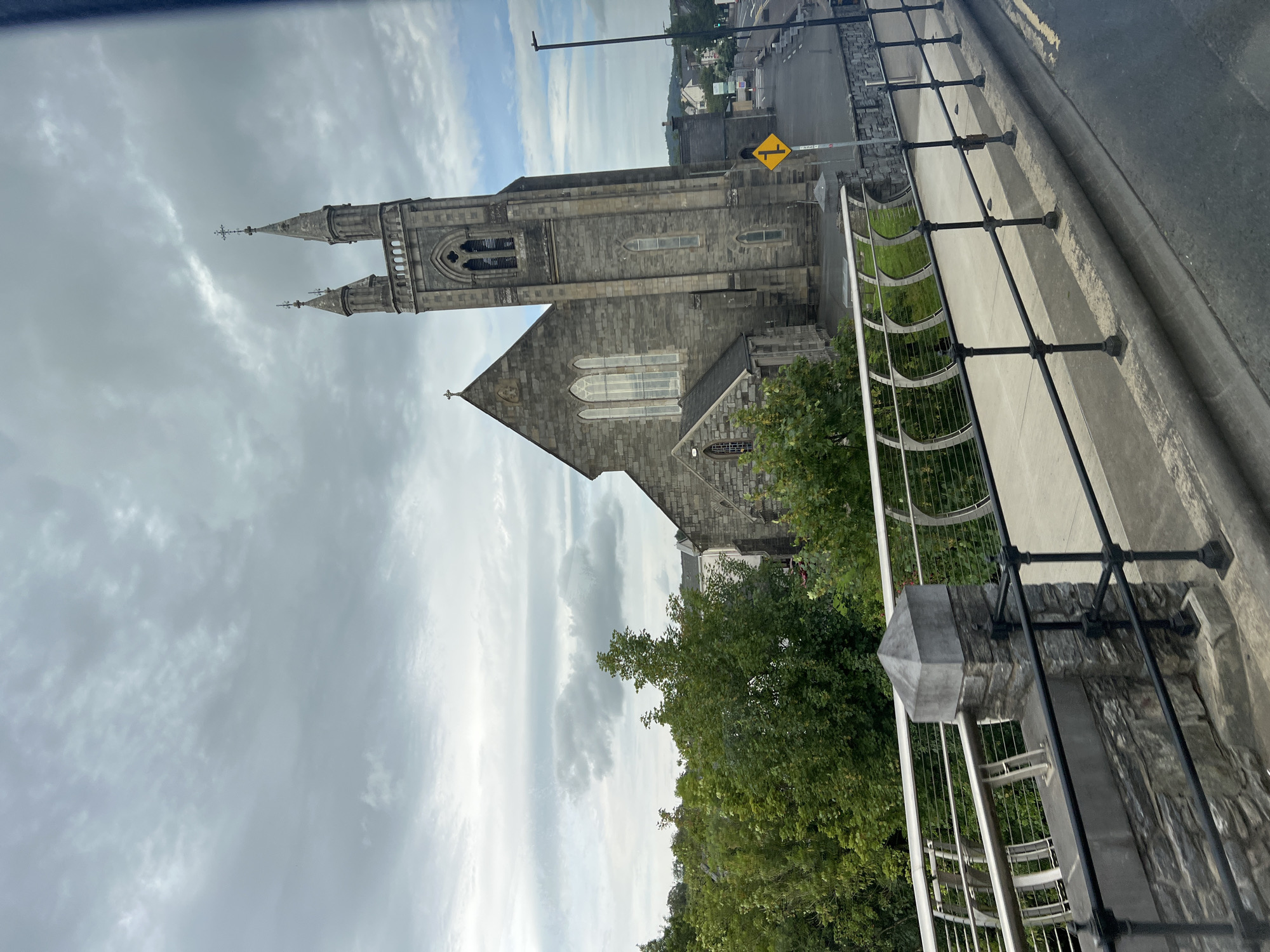 The image captures a view of St. Mary's Church in Ballybofey, County Donegal, Ireland, as seen from a bridge. The church is a prominent stone structure with a tall tower featuring spires and crosses, a pitched roof, and arched windows. Adjacent to the church are lush green trees. In the foreground, a pedestrian walkway is visible, bordered by a modern railing system made of stone and metal. The walkway leads to a street, with a yellow diamond-shaped traffic sign for a right turn visible. The sky is overcast with a mix of clouds. The scene suggests a partly cloudy day.
