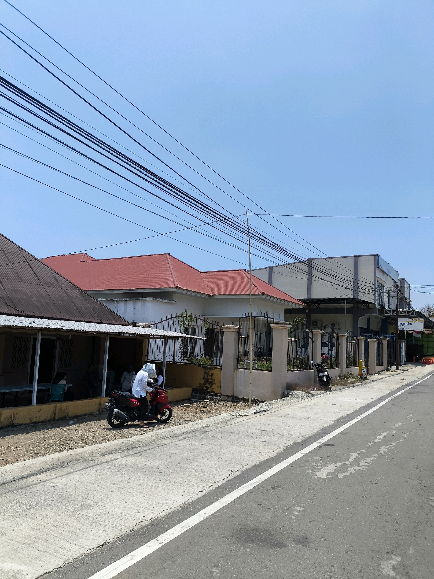 A street scene in Kwandang, Indonesia, shows a paved road with a white line marking the lane. On the left side of the road, there are several buildings. The closest building on the left has a corrugated metal roof and an open front with people seated inside. Next to it is a building with a red tiled roof. Further down the street, a modern building with a flat roof and gray and white facade is visible, featuring signage for "mandiri." Power lines are strung across the sky above the buildings. A red motorcycle is parked on the side of the road, with a person in a white shirt and head covering sitting on it. A black motorcycle is parked further down near a fence. The ground beside the road is covered with gravel and some patches of grass. The sky is clear and blue.