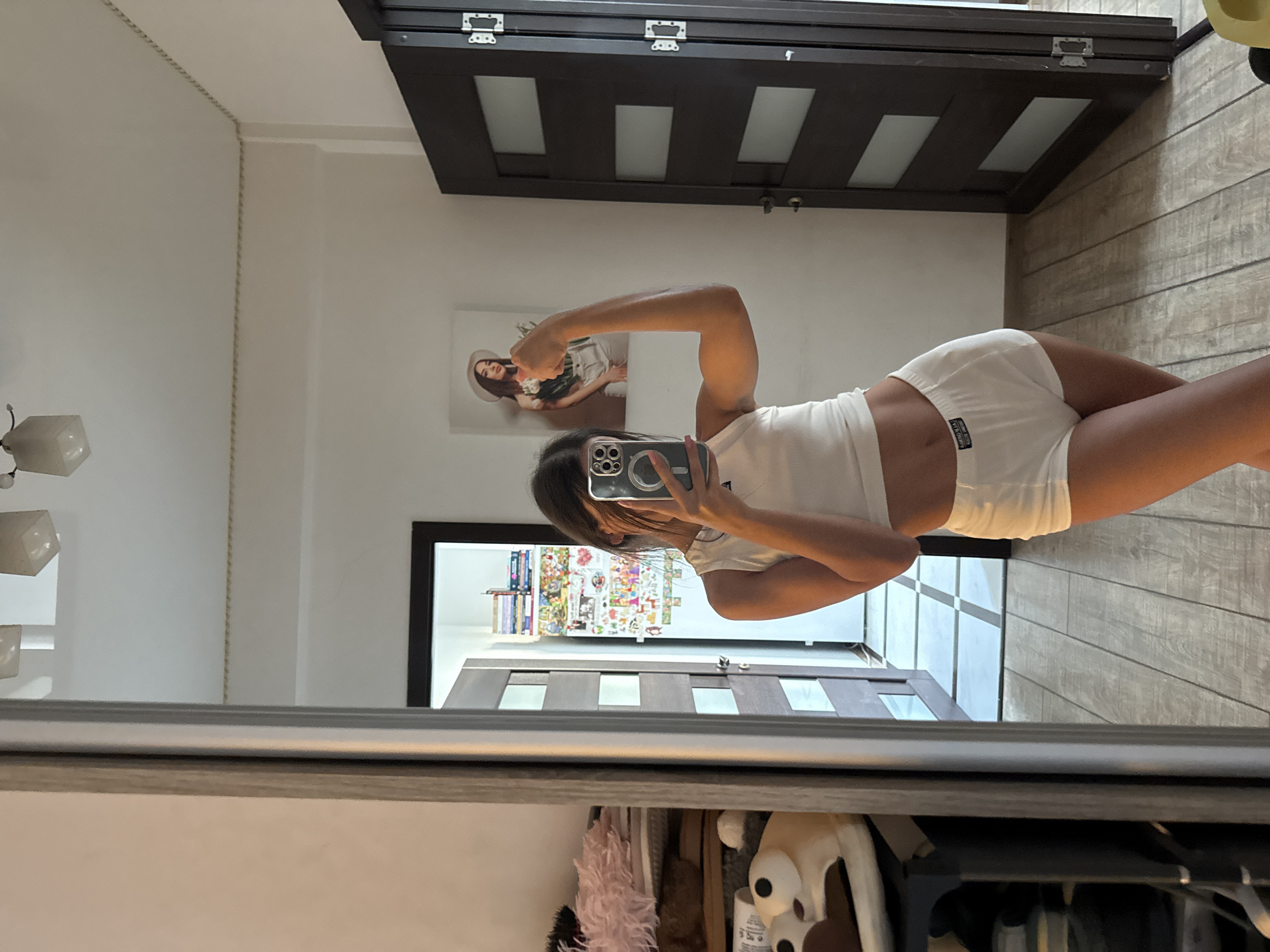 This is a vertical, eye-level, medium shot of a person posing for a mirror selfie. The subject is a woman with long dark hair, wearing a white tank top and white shorts. She is flexing her bicep, and the camera is held in her right hand, obscuring her face. The mirror reflects a room with wooden floors and a dark brown door with three rectangular glass panes. There is a framed picture on the wall behind the subject, depicting a woman wearing a hat and holding flowers. In the reflection of the mirror on the opposite wall, a closet is visible with several shelves filled with colorful items, possibly books or artwork, and a dark-colored door. A floor lamp with a white square shade is also visible in the upper left corner of the frame. The lighting suggests it is daytime, and the setting appears to be a residential interior. No specific weather conditions or emotions are evident.
