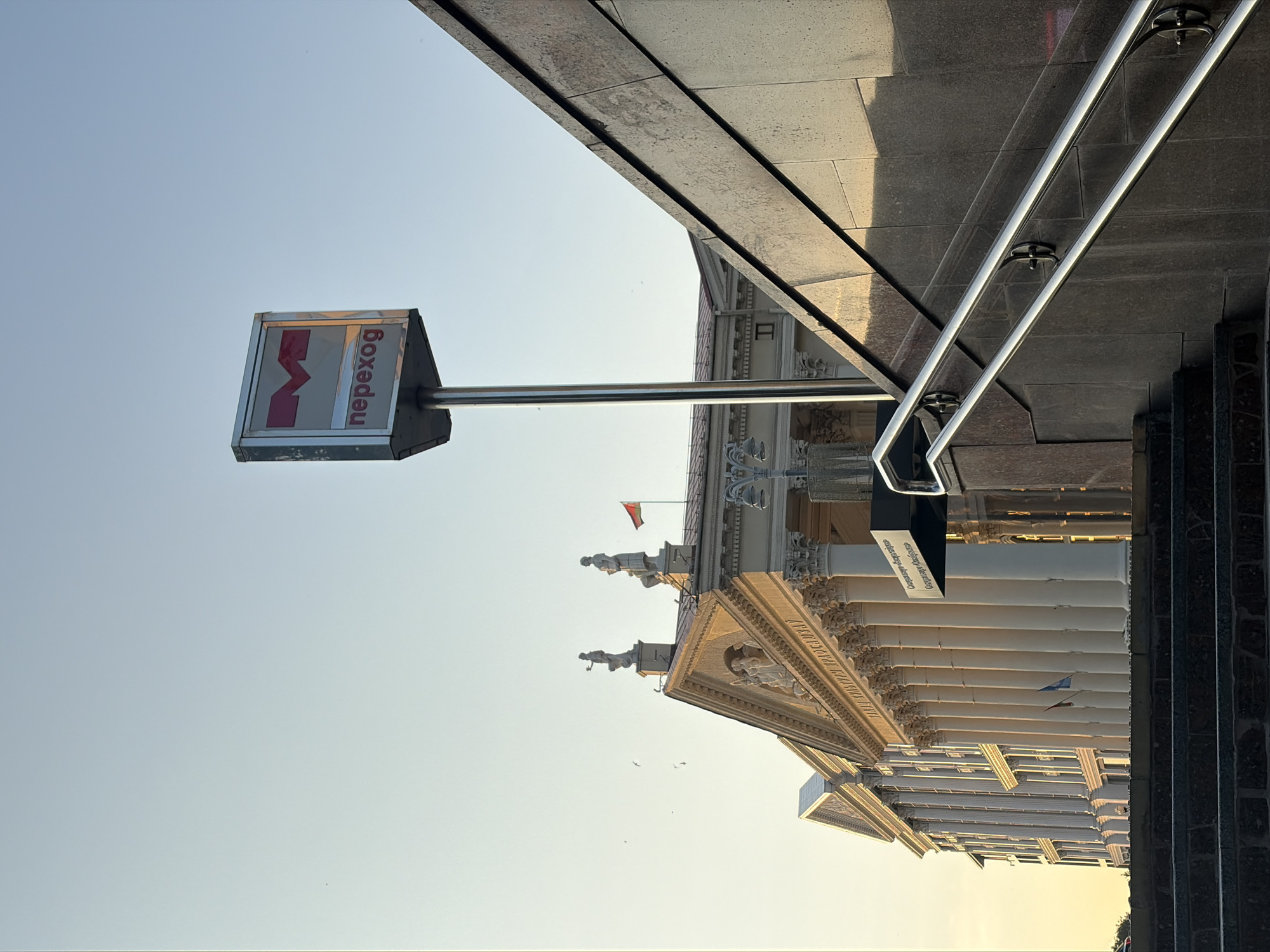 The image shows the entrance to a subway station in Minsk, Belarus. The entrance is marked by a prominent sign with a red "M" logo and the word "переход" (perekhod), meaning "transfer" or "underpass" in Russian. The sign is mounted on a tall metal pole. To the right of the sign, a modern granite staircase with a stainless steel handrail ascends. In the background, a large neoclassical building with rows of columns and statues can be seen, suggesting it might be a significant public building or institution. The sky is clear and bright, indicating it is daytime, likely during the golden hour due to the warm light casting shadows and highlighting the architecture. There are no people visible in the frame, and the scene is still. The overall impression is one of urban infrastructure against a backdrop of historical architecture.