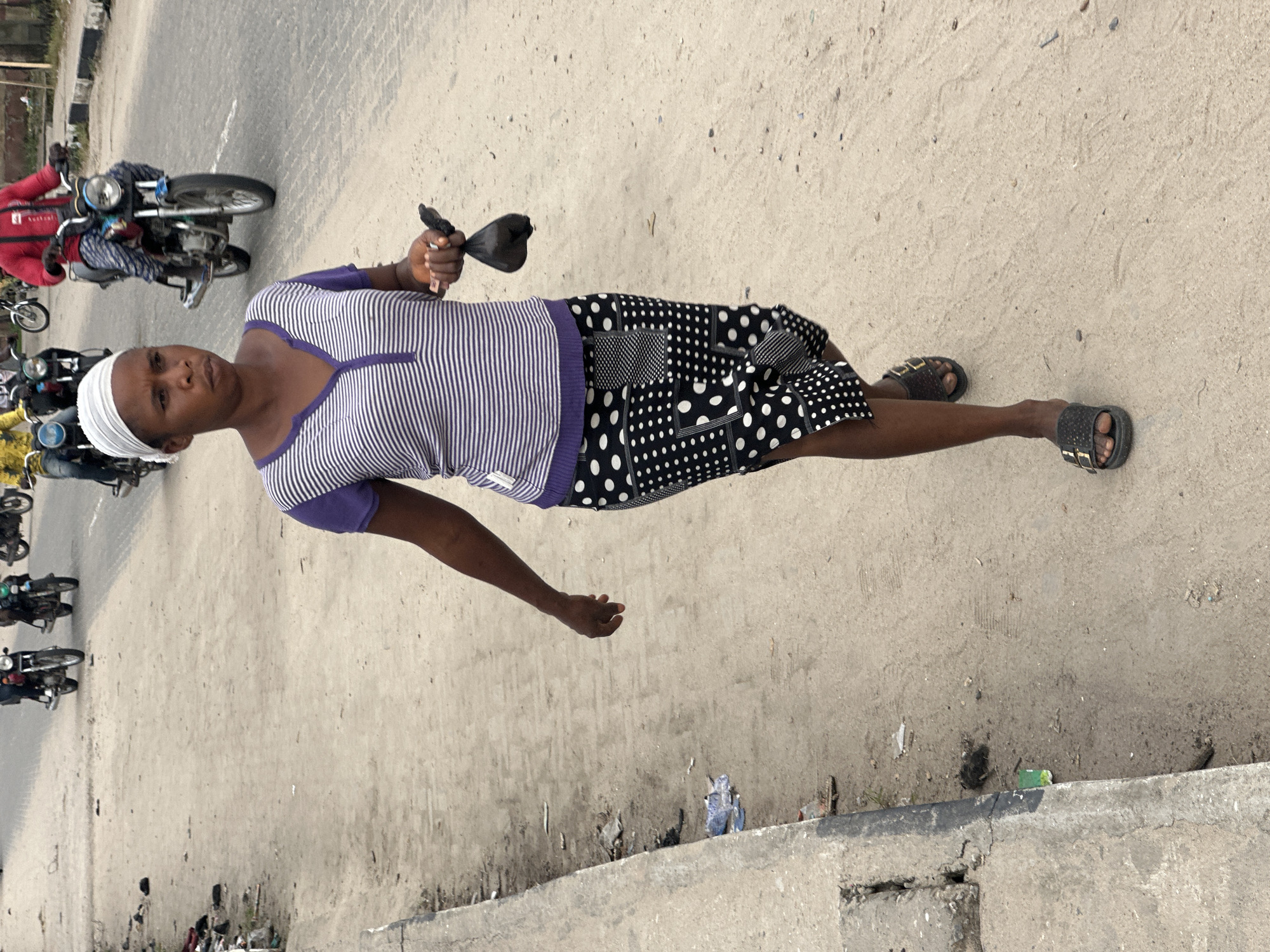 In Badagry, Nigeria, a woman walks along a dirt road beside a concrete curb. She is wearing a striped shirt, a patterned skirt, and sandals, and carries a black bag in her right hand. In the background, several motorcycles are visible on the road, with riders on them. The road is unpaved, with some debris scattered along the sides. The environment appears to be an urban or semi-urban area.