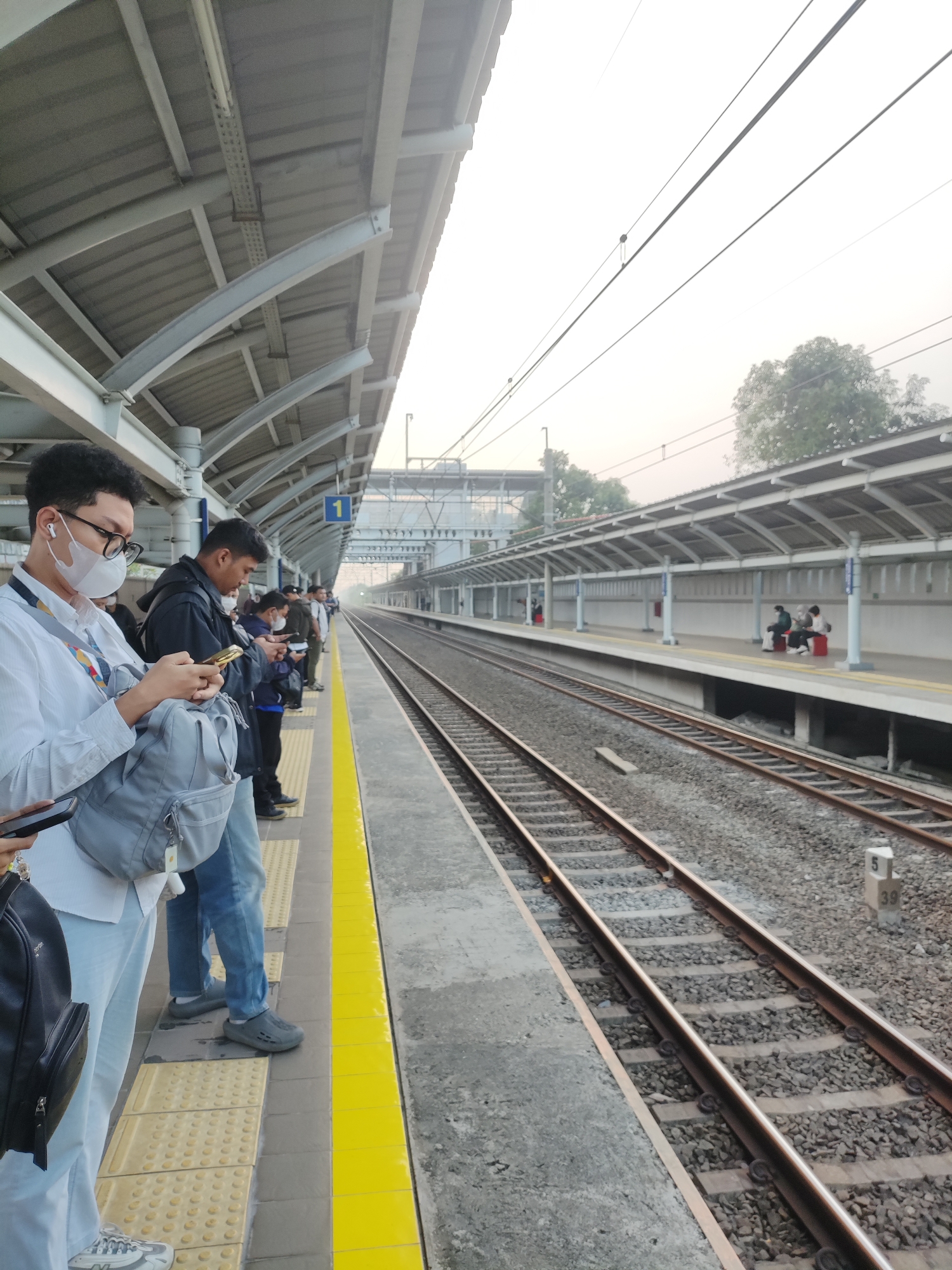 The image depicts a train station platform in Telagamurni, Indonesia, with several people waiting for a train. The platform has a covered roof supported by grey metal beams and pillars. Tracks run alongside the platform. Several individuals are standing and looking at their mobile phones. A person in the foreground wears glasses, a white shirt, a face mask, and AirPods, and carries a grey bag. Another person in a black jacket is also focused on their phone. Further down the platform, other people are visible, some seated on red benches. The environment appears to be an outdoor setting with neutral lighting, suggesting it could be morning or late afternoon. The ground of the platform is made of concrete with yellow tactile paving near the edge. The overall scene is one of quiet waiting at a public transportation hub.