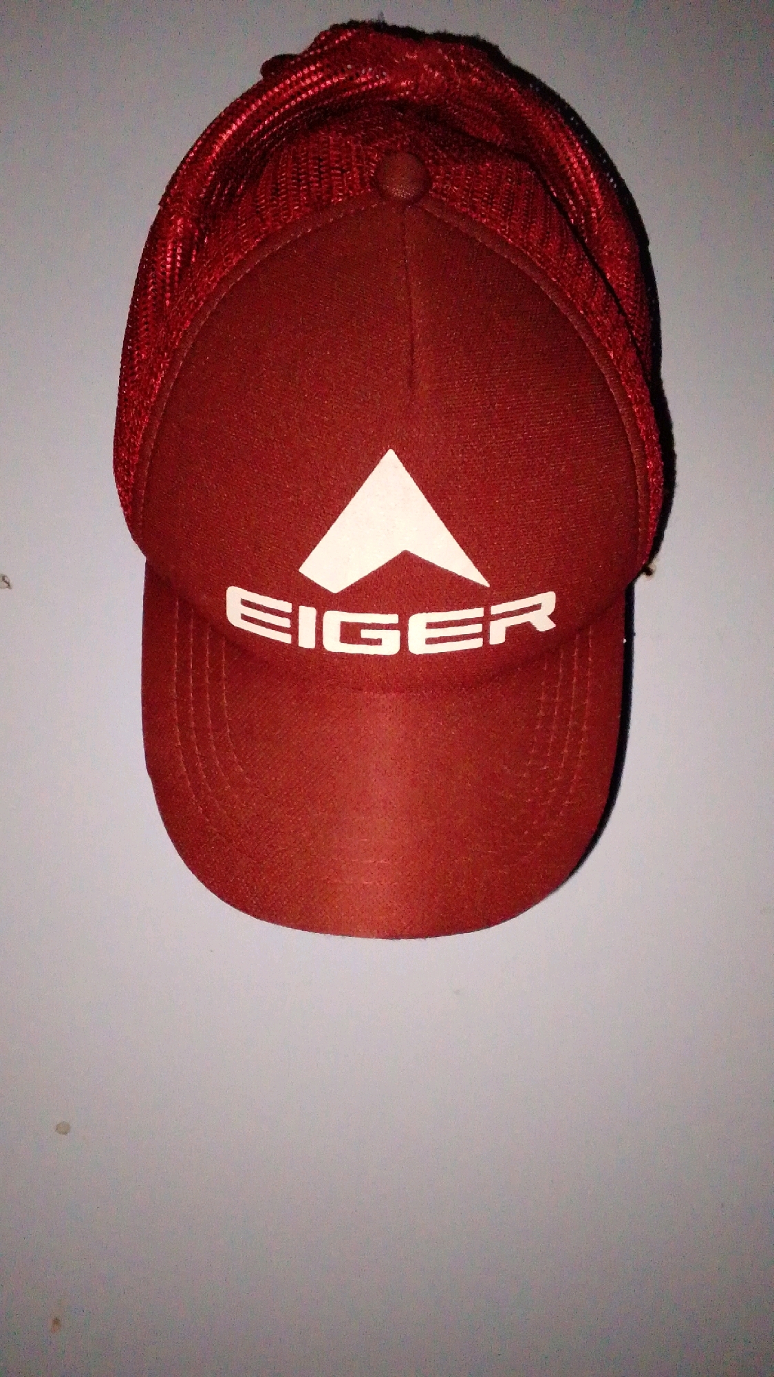 A red baseball cap with a white logo is displayed against a light gray wall. The logo features a white triangle above the word "EIGER" in white block letters. The cap has a mesh back and a structured front panel. The background wall appears to be textured and has some minor imperfections, possibly smudges or small marks. The lighting suggests an indoor setting. No specific location in Cintaratu, Indonesia, can be determined from the image.
