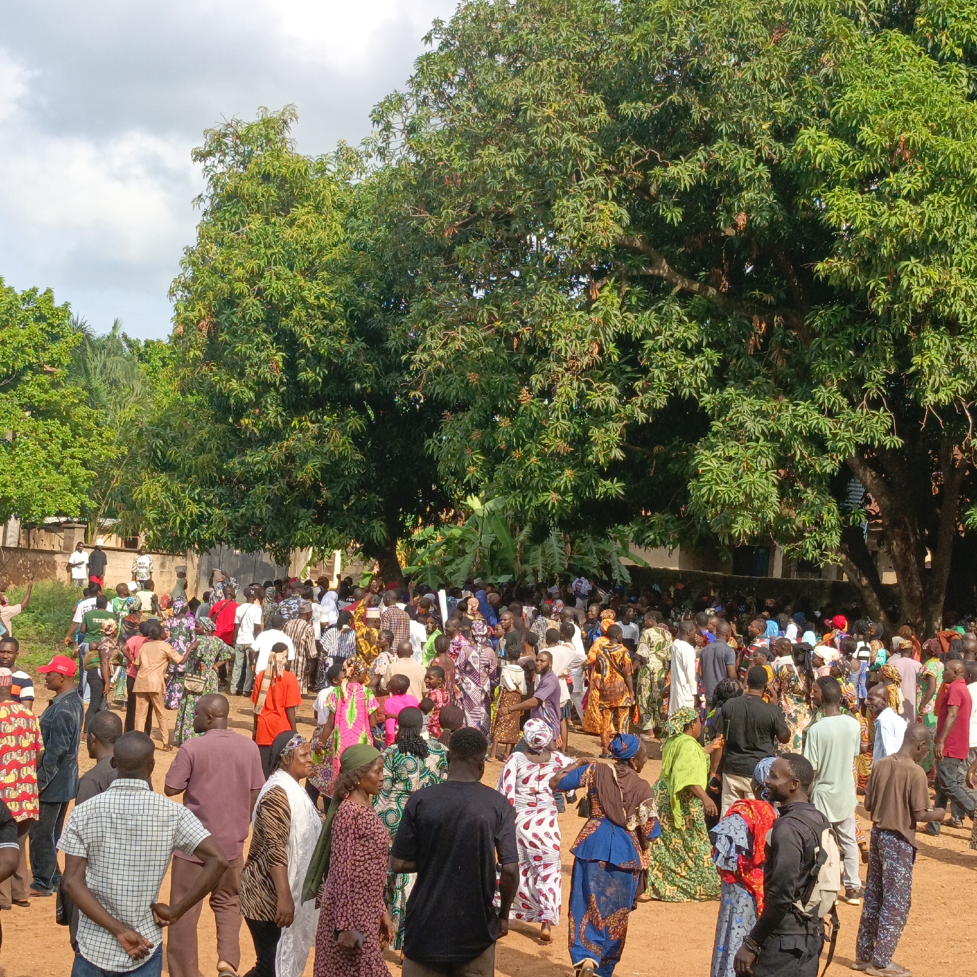 The image depicts a large gathering of people in Akwanga, Nigeria. The crowd is diverse, with individuals wearing a variety of clothing styles, many in colorful traditional fabrics. A large tree dominates the background, providing shade. The ground is reddish-brown, suggesting dirt or clay soil. The environment appears to be outdoors, possibly a village square or community gathering space. Actions and interactions are not clear.