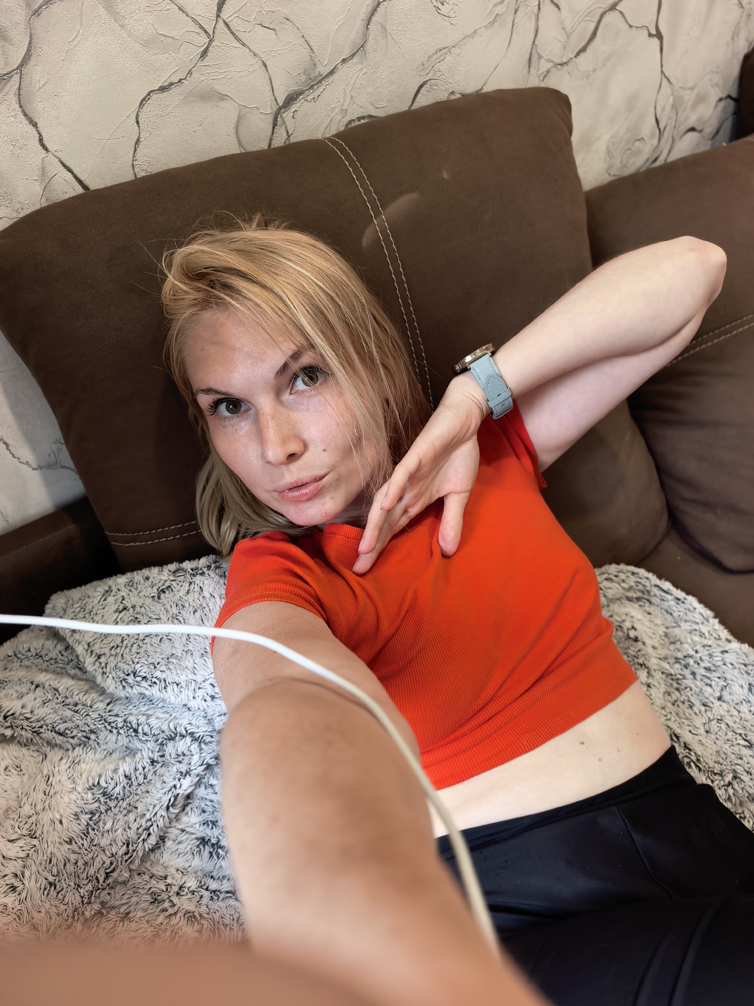 A blonde woman lies on a brown couch covered by a soft, grey and white blanket. She is wearing an orange top and black pants, with a light blue watch on her left wrist. A white cable runs across the frame, partially obscuring her. The couch is positioned against a textured wall with a marble-like pattern. The scene appears to be indoors, likely in a residential setting within the city of Sterlitamak, Russia.