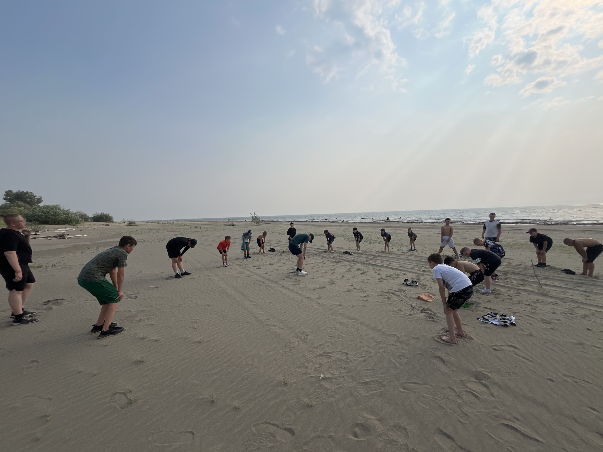 A group of young men are gathered on a sandy beach, seemingly engaged in a warm-up or stretching routine. The scene takes place outdoors, with a calm body of water, likely a lake or sea, stretching to the horizon under a hazy sky. The sun is low, suggesting it's either early morning or late afternoon, casting a soft light and creating long shadows on the sand. The weather appears to be warm and somewhat overcast, with scattered clouds.
The subjects are dressed in athletic wear, including t-shirts, shorts, and sneakers. They are positioned in a line or semi-circle, with many bending over or stretching their bodies. Their postures indicate a focus on physical activity. There are no discernible objects other than their clothing and some scattered shoes and a small orange shovel. The ground is covered in fine sand with some sparse vegetation growing in patches. The background shows more sandy terrain and distant, hazy treelines. The overall mood appears to be one of shared physical exertion and possibly training. There is no visible text in the image.