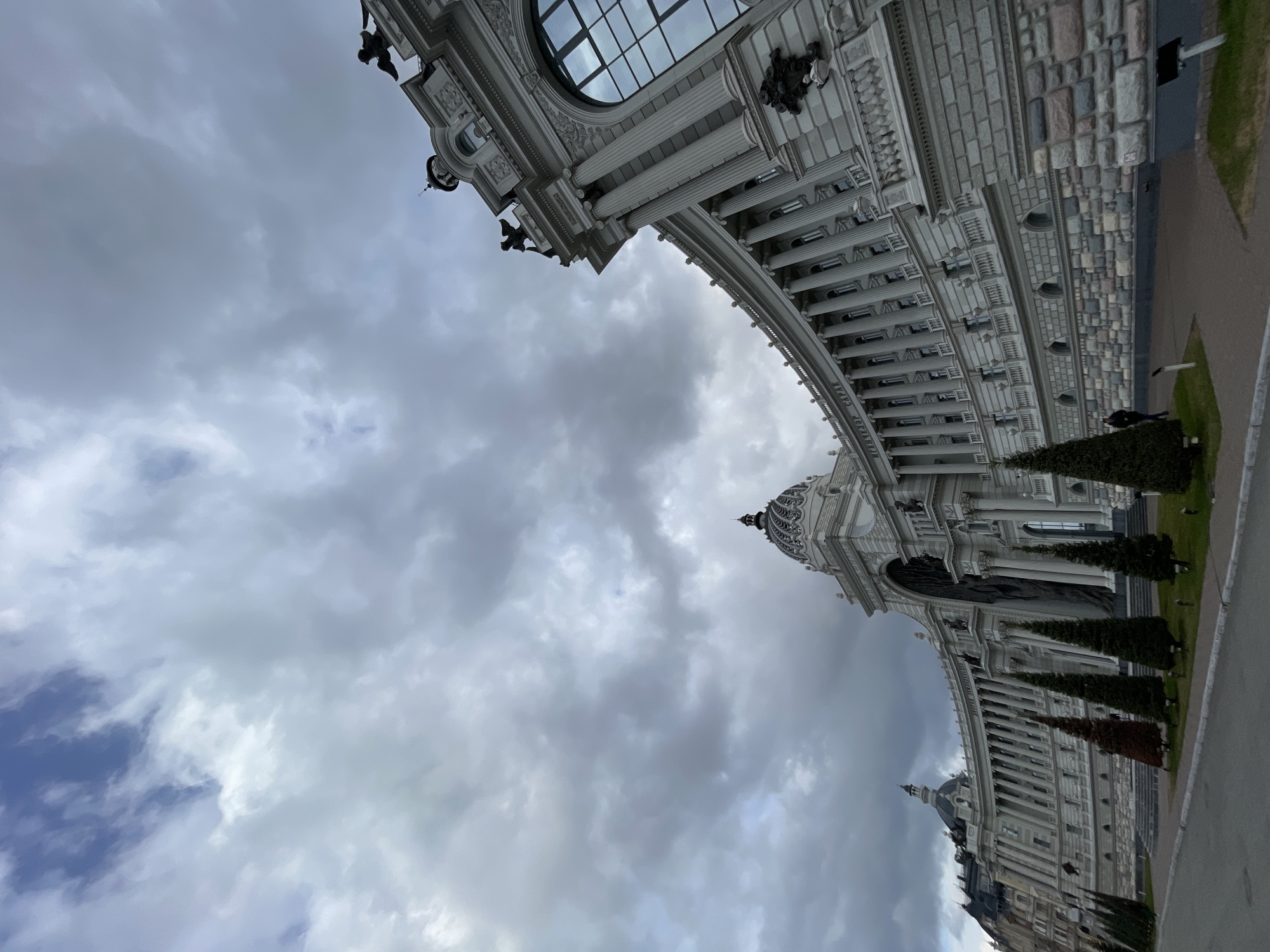 The image captures a grand, ornate building showcasing classical architectural elements under an overcast sky. The structure, rendered in light-colored stone or concrete, occupies the upper and right portions of the frame, viewed from a low, upward-looking perspective.
The building features a prominent arched passage, numerous fluted columns, and intricate decorative friezes and carvings. A large, semi-circular window or skylight with a grid pattern is visible high on the facade. Further along the building's curving expanse, a large, intricately patterned dome and a smaller cupola can be seen. Decorative figures, resembling gargoyles or statues, are placed on various ledges. The facade displays a mix of smooth panels, textured block-like masonry, and highly detailed sculptural work. Partially visible text on an archway reads "ВЕНЦОВЫЙ ДВОРЕЦ," which translates to "Crown Palace" or "Wedding Palace."
The sky fills a substantial portion of the image, dominated by dense, grey clouds, indicating an overcast daytime. Some areas of the clouds are lighter, and a small patch of blue sky is visible towards the bottom left.
Along the lower right edge, a narrow strip of green grass borders a grey paved path or road. Several conical evergreen trees or shrubs are neatly arranged along the building's base. A solitary, indistinct person is faintly visible near the building's foundation in the distance.
The scene conveys architectural majesty and scale, with the cloudy weather adding a dramatic or somber mood. The building is identifiable as the Agricultural Palace (Dvorets Zemledeltsev) in Kazan, Russia, aligning with the provided location context.