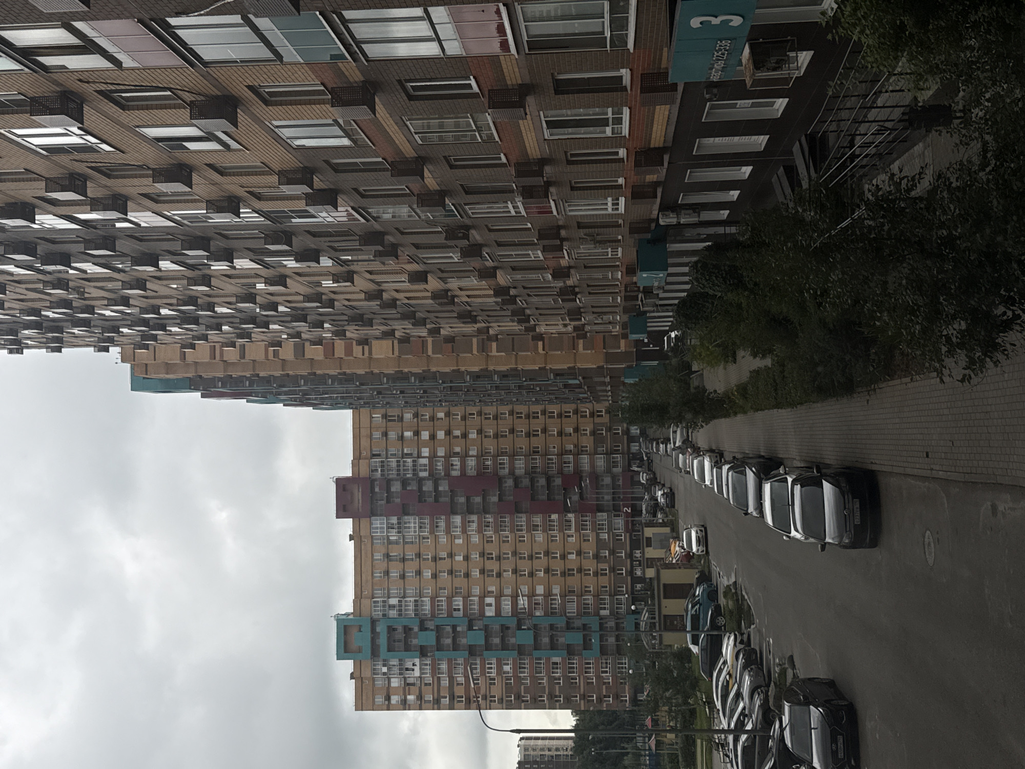 The image shows a street scene in Misaylovo, Russia, featuring a row of modern, multi-story apartment buildings and parked cars. The buildings are tall and have a contemporary design, with a mix of brick and colored facades. The apartment buildings are densely packed, suggesting an urban or suburban residential area. Several cars are parked along the street and in a parking lot, indicating a typical day in a residential neighborhood. The sky is overcast, casting a muted light on the scene. The environment appears to be a well-maintained urban area, with sidewalks and some greenery. There are no people visible in the image.
