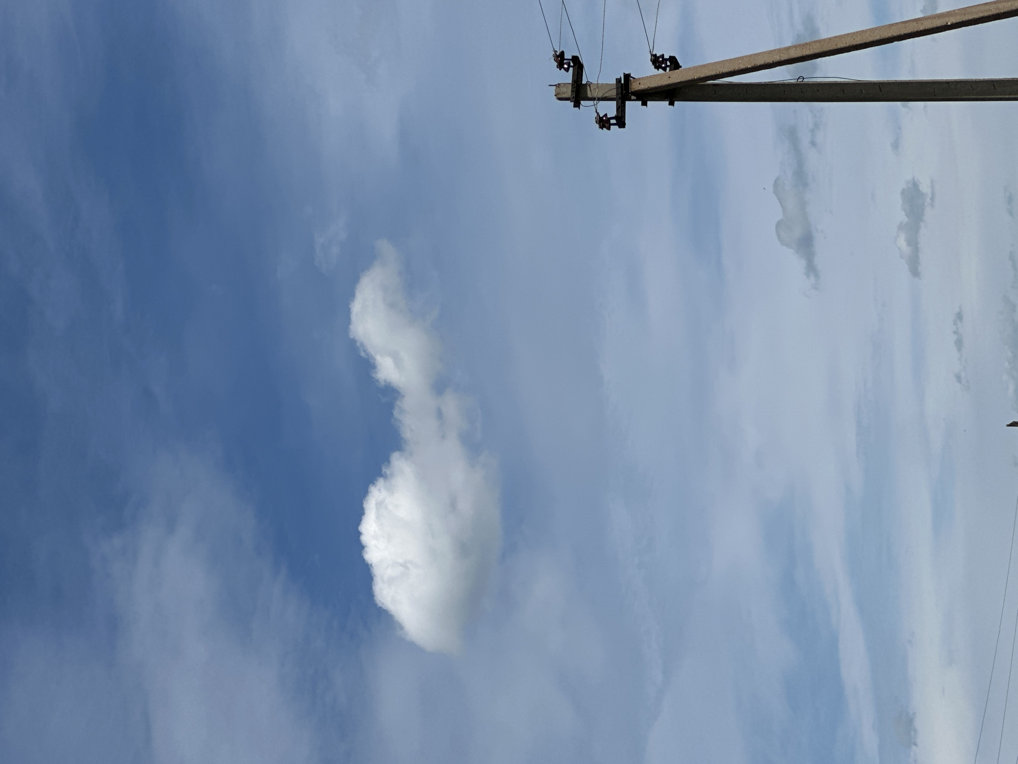 A concrete utility pole stands on the right side of the frame, its wires stretching across the image. The sky is a mix of blue and white clouds. A large, puffy white cloud is visible in the upper left quadrant, appearing to be the most prominent cloud formation in the image. Several other, smaller clouds are scattered across the sky. The lower portion of the image shows the base of the utility pole and a faint hint of what appears to be power lines extending into the distance. The scene is devoid of any visible people or animals. The overall environment suggests an outdoor setting under a partly cloudy sky.