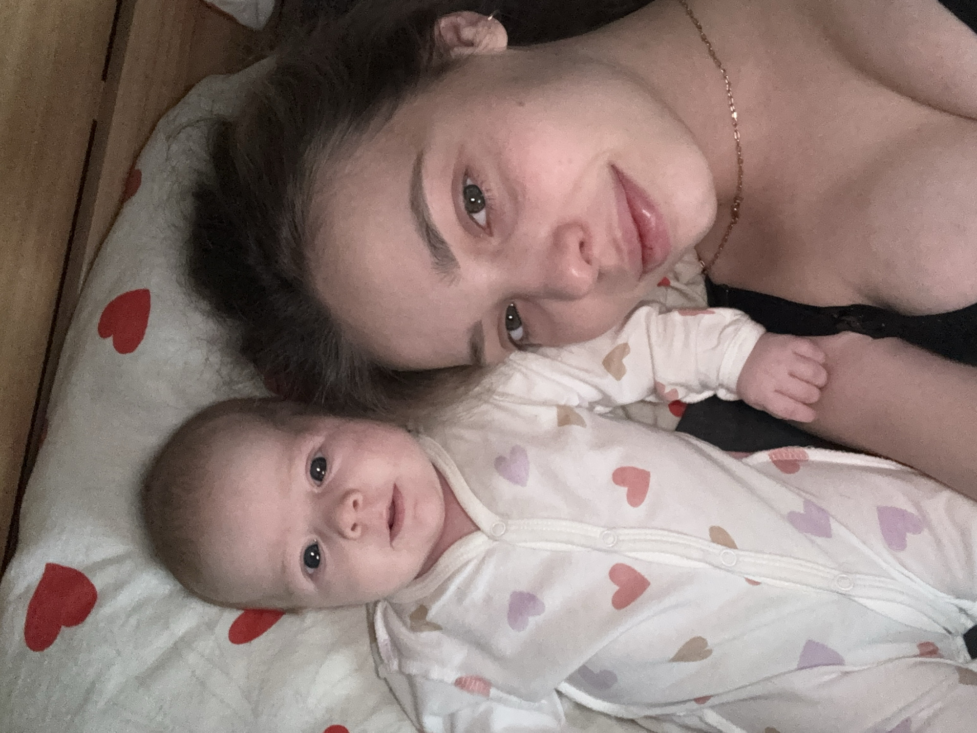A woman and an infant lie on a heart-patterned bedsheet. The woman has brown hair and light skin, and she is wearing a gold necklace and a black top. The infant is wearing a white onesie with pink and purple hearts. Both are looking directly at the camera. The setting appears to be indoors, potentially a home in the city of Chaikovsky, Russia.