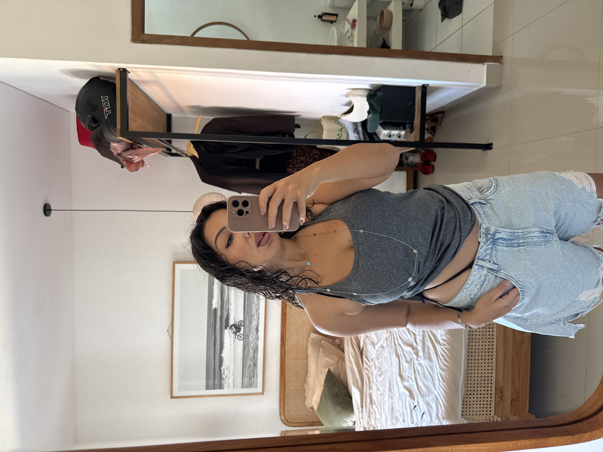 This is a vertical, eye-level, medium shot captured in what appears to be a bedroom in Kuta Selatan, Indonesia. The primary subject is a woman taking a mirror selfie, with her phone prominently featured. She is wearing a grey tank top and light-wash denim shorts with ripped detailing. A delicate silver necklace with a blue pendant is visible around her neck. Her wet, dark hair frames her face as she sticks out her tongue playfully.
The background reflected in the mirror reveals a well-lit room with white walls. A large framed black and white photograph of a motorcycle on a beach hangs on the wall. To the right, a bed with white and green linens is visible, with a headboard made of woven rattan. A wooden shelf unit is mounted on the wall, holding a black helmet and various clothing items, including what appears to be a patterned scarf. The overall atmosphere suggests a casual, relaxed environment, possibly a hotel room or a personal residence. There is no indication of the time of day or weather conditions. The only visible text is on the helmet in the background, which reads "TOI."