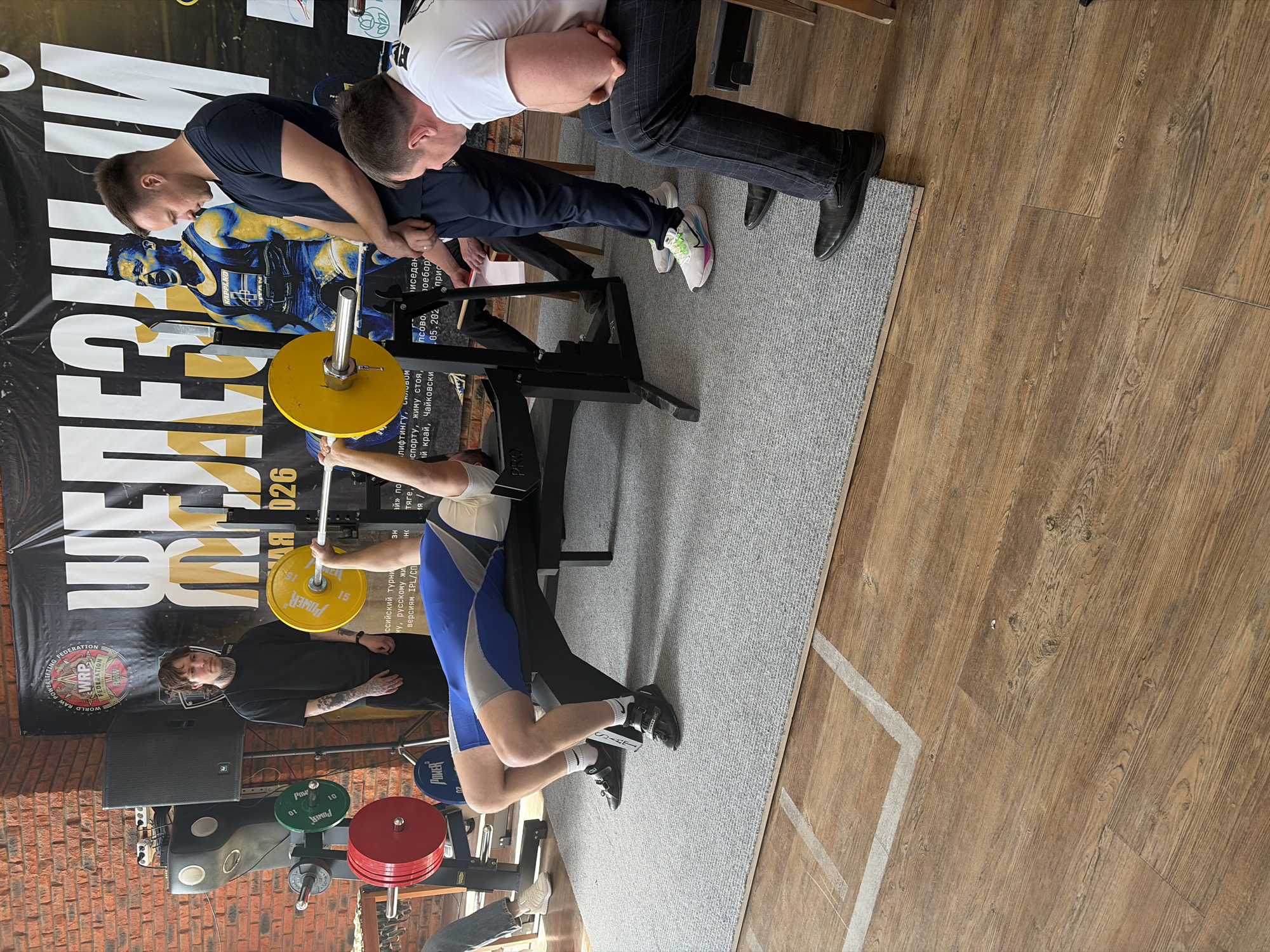 The image shows a man performing a bench press exercise, assisted by two spotters. The barbell is loaded with yellow and other weights. Behind them, there is a poster with text in Russian, and logos for a powerlifting federation and a local tournament. A man stands near a speaker watching the event, revealing tattoos on his arm. In the background, there's a brick wall. The scene appears to be taking place at a powerlifting competition or gym in the city of Chaikovsky, Russia, as suggested by the Russian text on the poster. The floor is wooden, and there is a grey rug under the bench press.