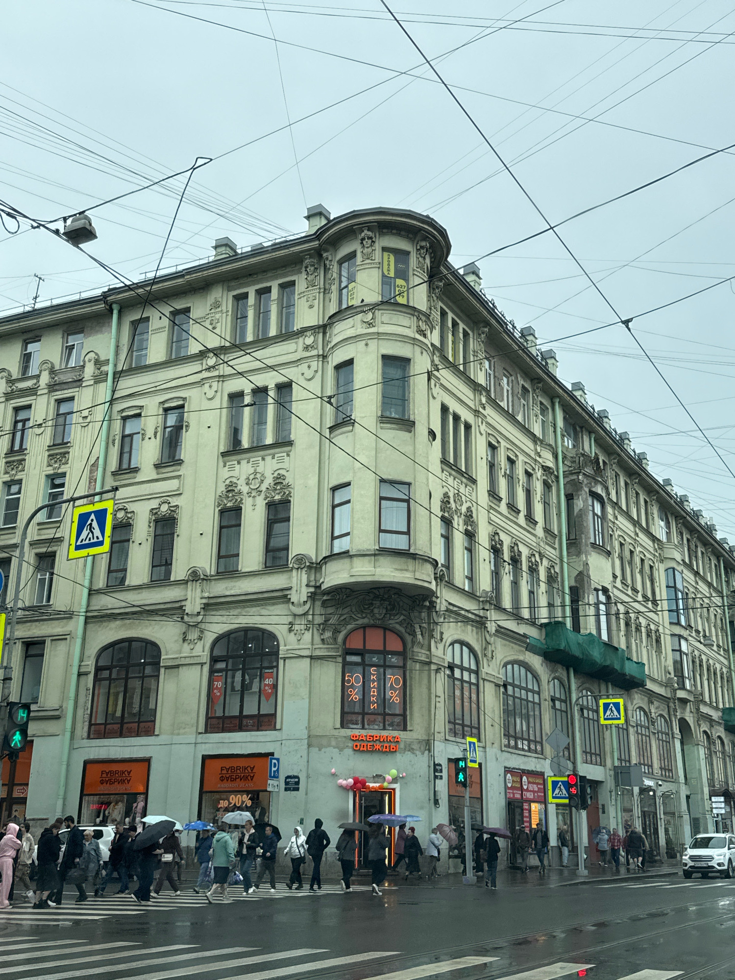 The image depicts a street scene in Saint Petersburg, Russia, on a cloudy day. The foreground shows a wet crosswalk with several people holding umbrellas and crossing the street. They are dressed in casual clothing, with some individuals wearing raincoats. A white car is visible in the distance on the right side of the frame.
The mid-ground features the facade of a multi-story building with light-colored walls and numerous windows. The architecture appears to be historic, with decorative elements on the exterior. Several storefronts are visible on the ground floor, including "FABRIKI ФУБРИКИ" and "ОДЕЖДЫ," indicating clothing stores. There are also advertisements displayed in the windows, some with percentages like "70 %" and "50 %". Traffic lights and street signs, including a pedestrian crossing sign, are present. Overhead, a complex network of electrical wires crisscrosses the sky.
The background is dominated by the overcast sky. The overall impression is of a typical urban environment in a European city, with the presence of rain and pedestrian activity suggesting a moderately busy day.