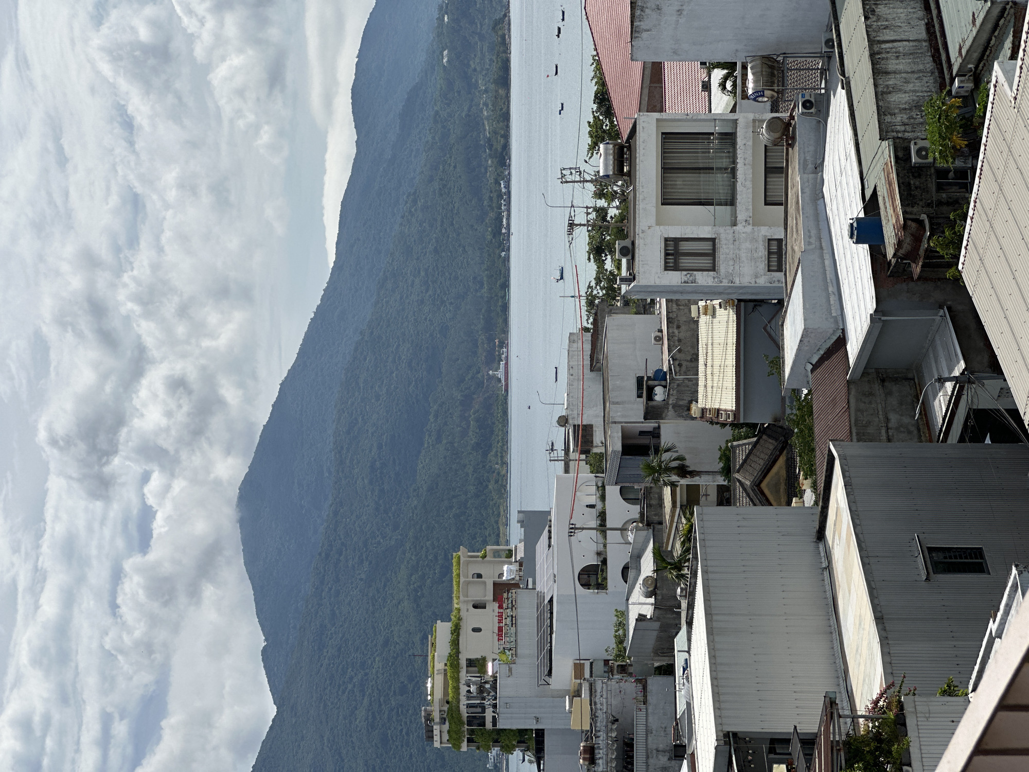 This is a high-angle, medium shot of a coastal city in Da Nang, Vietnam, looking towards a bay with mountains in the background. The foreground is dominated by a dense cluster of residential and commercial buildings with various roof types, including corrugated metal and flat concrete. Many of the buildings are white or light-colored, some with visible air conditioning units and water tanks on their roofs. Solar panels are visible on at least one building.
In the middle ground, the bay stretches out, dotted with several small fishing boats and one larger vessel in the distance. The water appears calm.
The background features a large, tree-covered mountain range that slopes down to the bay. The sky is partially overcast with fluffy, white clouds, suggesting either morning or late afternoon. There are no visible people or animals in the frame. The scene depicts a typical urban coastal environment in Vietnam, with a blend of everyday buildings and natural scenery.