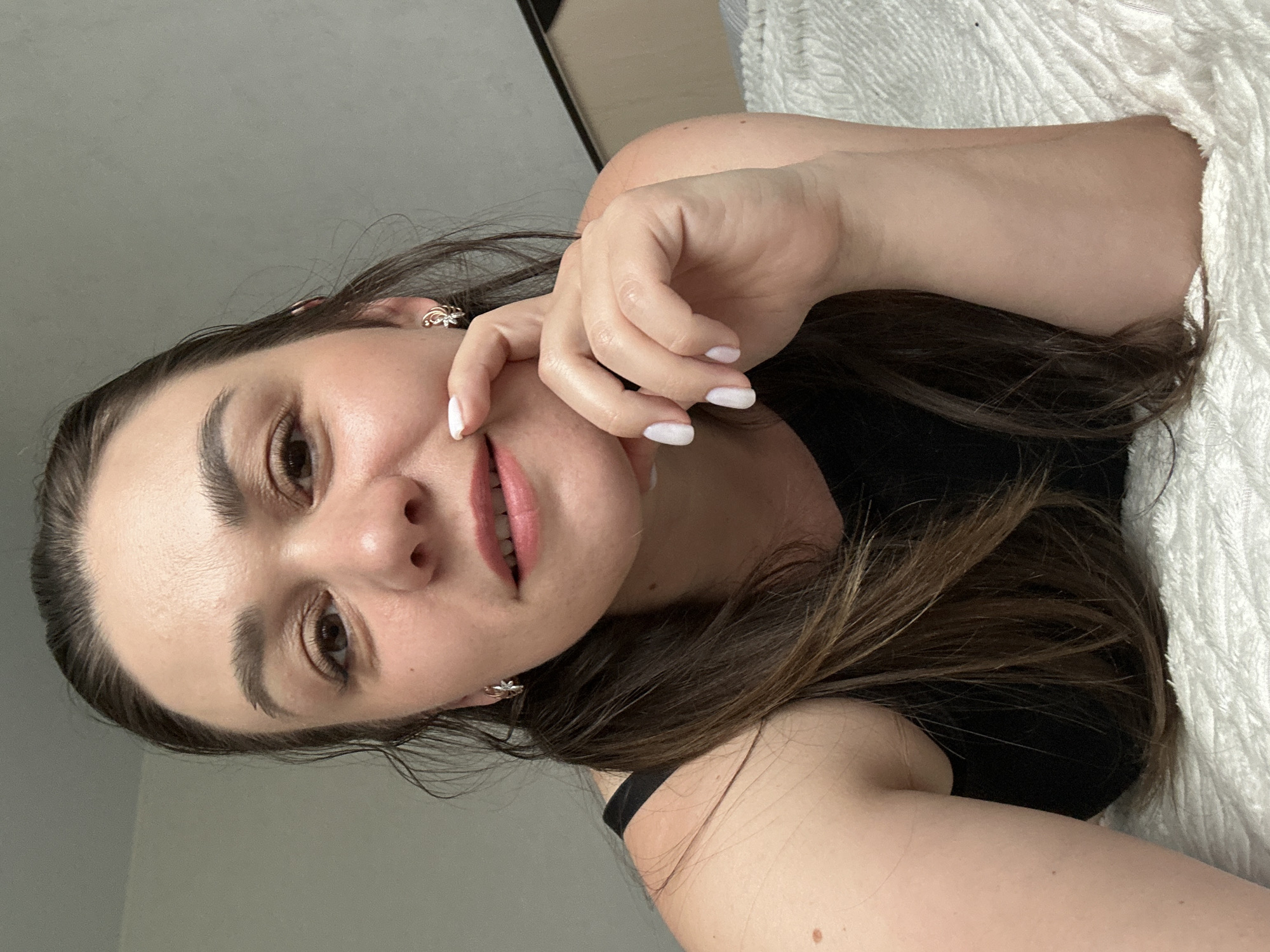 A woman with brown hair is lying on a white textured surface, possibly a bed. She is wearing a black top. Her right hand is touching her face. She is wearing small earrings. The background appears to be a grey wall. The photo was taken indoors. There is no information to determine the location is in Chaikovsky, Russia.