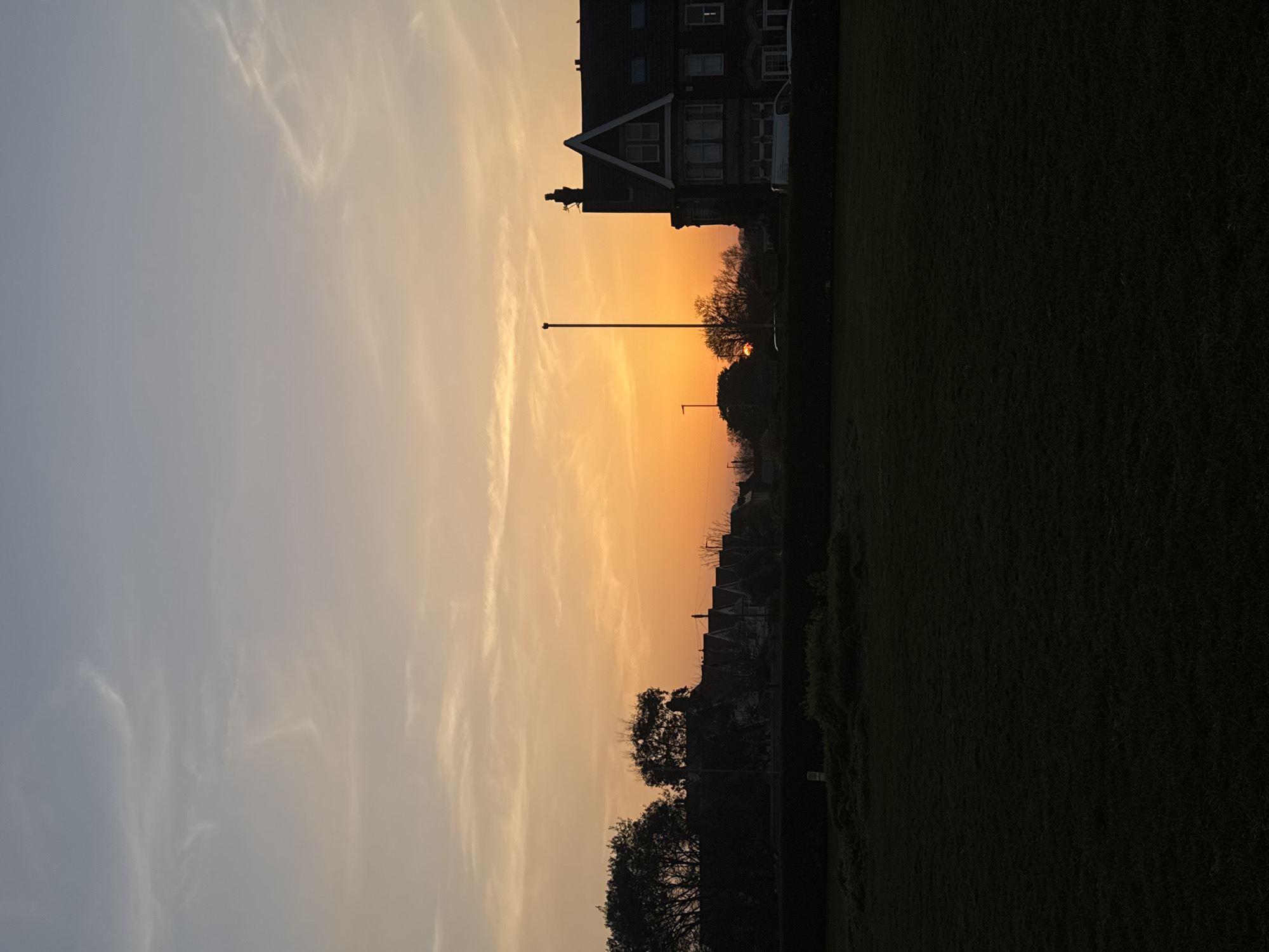The image depicts a landscape at dusk or dawn in Gorleston-on-Sea, United Kingdom. An orange-colored sky with faint clouds suggests either sunrise or sunset. A building with multiple windows and a pointed roof section is visible. Silhouetted trees and a lamp post are positioned in the foreground, and a dark green field is on the right side.