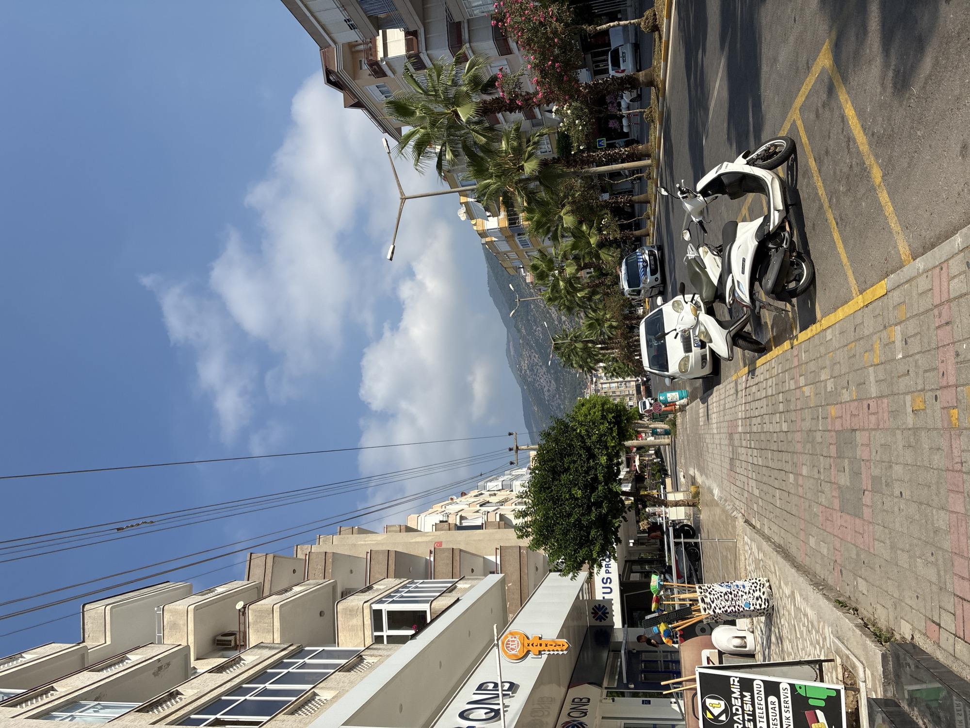 The image shows a street scene in Alanya, Turkey, with buildings and palm trees lining the road. The buildings are multi-story and appear to be apartments or commercial spaces. The street is paved and has yellow parking lines. Several white scooters and cars are parked along the street. There are power lines overhead, and the sky is blue with some clouds. In the foreground, there is a sidewalk with a store on the left, displaying a sign with the QNB logo and a large orange key-shaped sign. A display of brooms and cleaning supplies is also visible on the sidewalk. Further down the street, mountains can be seen in the distance. The overall impression is of a sunny day in a Mediterranean city.