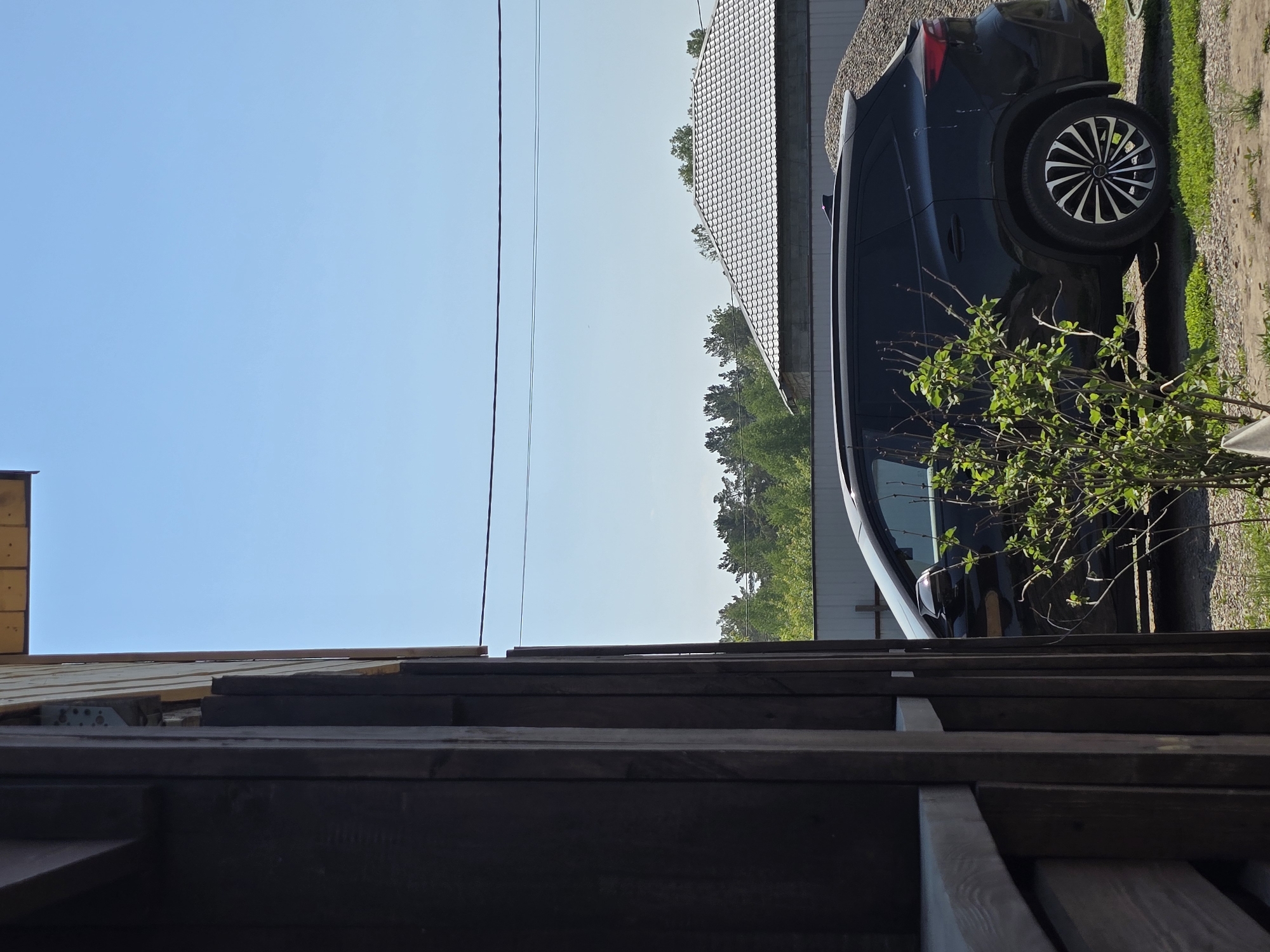 A forensic, matter-of-fact description of the scene in Ермолаевский Затон, Russia:
A daytime exterior shot captures a stationary black or dark-colored SUV, likely a crossover, parked on a gravel and patchy grass surface. The vehicle's right side is prominently displayed, featuring multi-spoke alloy wheels. In the immediate foreground, a small, leafy green bush partially obscures the front passenger side of the SUV. To the left, a dark brown wooden structure, possibly a building or fence, occupies the left third of the frame. In the mid-ground, behind the vehicle, a grey tiled roof with a distinct scalloped pattern is visible, belonging to a building constructed with light-colored concrete blocks. Adjacent to this building, a large pile of light-colored gravel or crushed stone is present. Further in the background, a line of green trees forms a natural boundary under a clear, cloudless blue sky. A single, thin black utility line stretches horizontally across the upper portion of the sky. No individuals are present in the scene.