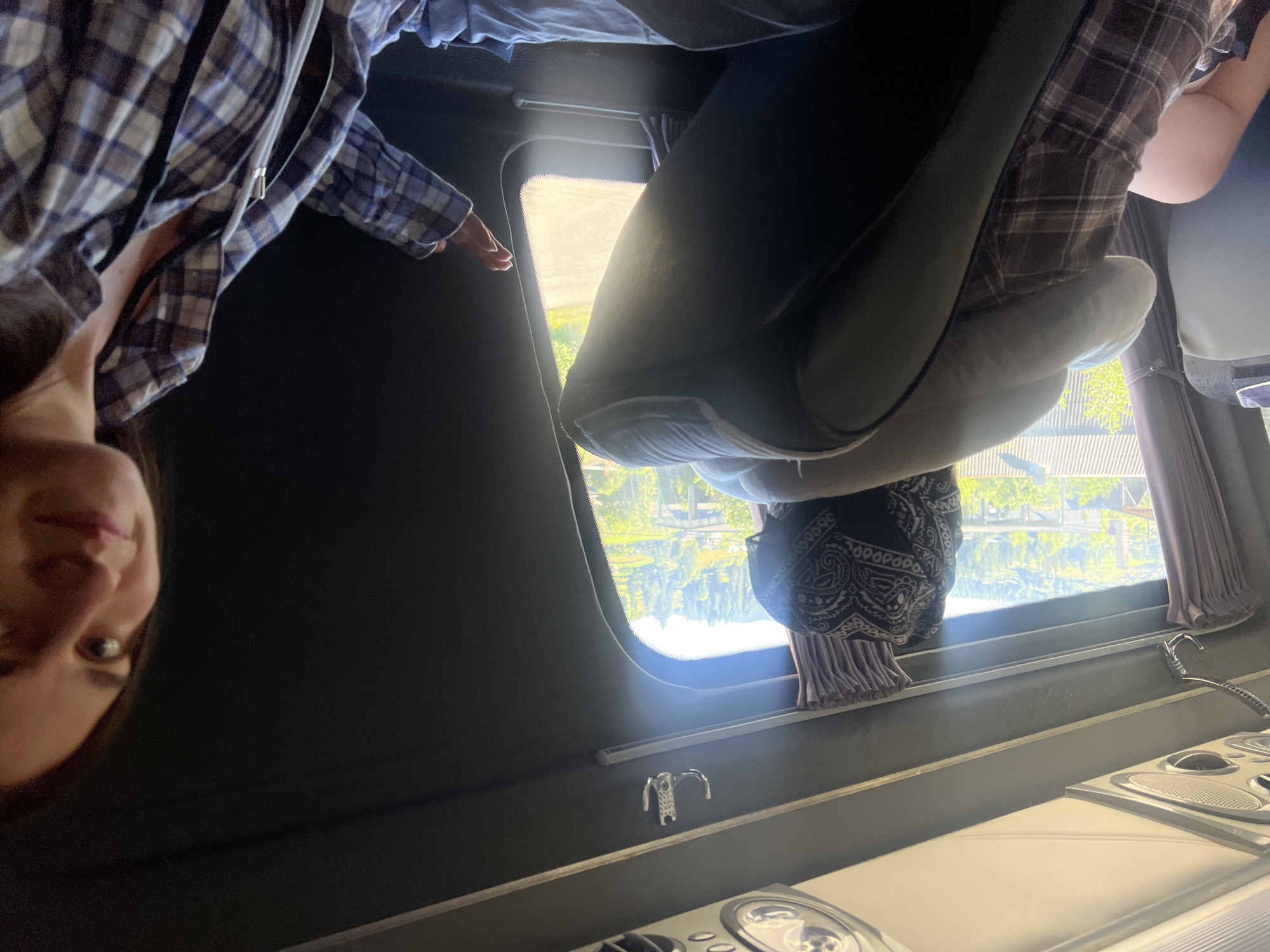 The image depicts the interior of a bus in Vorokhta, Ukraine, with two individuals. In the foreground on the right, a woman with long brown hair is wearing a blue and white plaid shirt and smiling. Her left hand is raised, appearing to wave or gesture. In the mid-ground, a person wearing a bandana and a plaid shirt is seated and leaning back against a pillow, looking out the window. The bus interior features dark walls, curtains, and a hook attached to the ceiling. Through the window, a lush green landscape with trees and buildings is visible under a bright sky, suggesting a daytime scene.