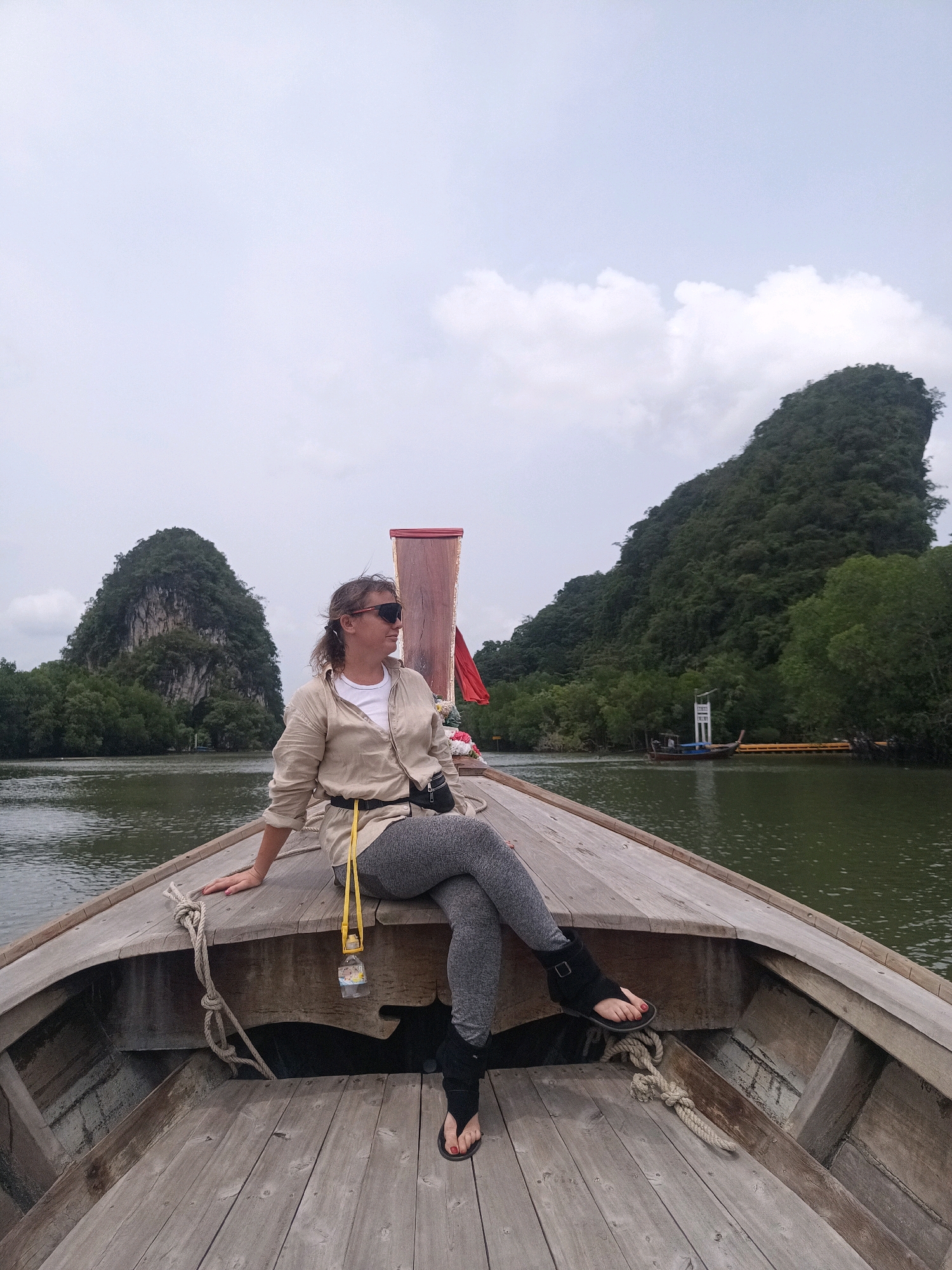 A woman sits on the bow of a wooden longtail boat in Khlong Prasong, Thailand. She is wearing a light-colored open button-down shirt over a white t-shirt, gray leggings, and black sandals. She has sunglasses on and her hair is pulled back. A yellow lanyard with a small water bottle hangs from her waist. The boat is on a body of water, with lush green karst mountains in the background. The sky is overcast. Another longtail boat is visible in the distance. The wooden planks of the boat are weathered and worn.