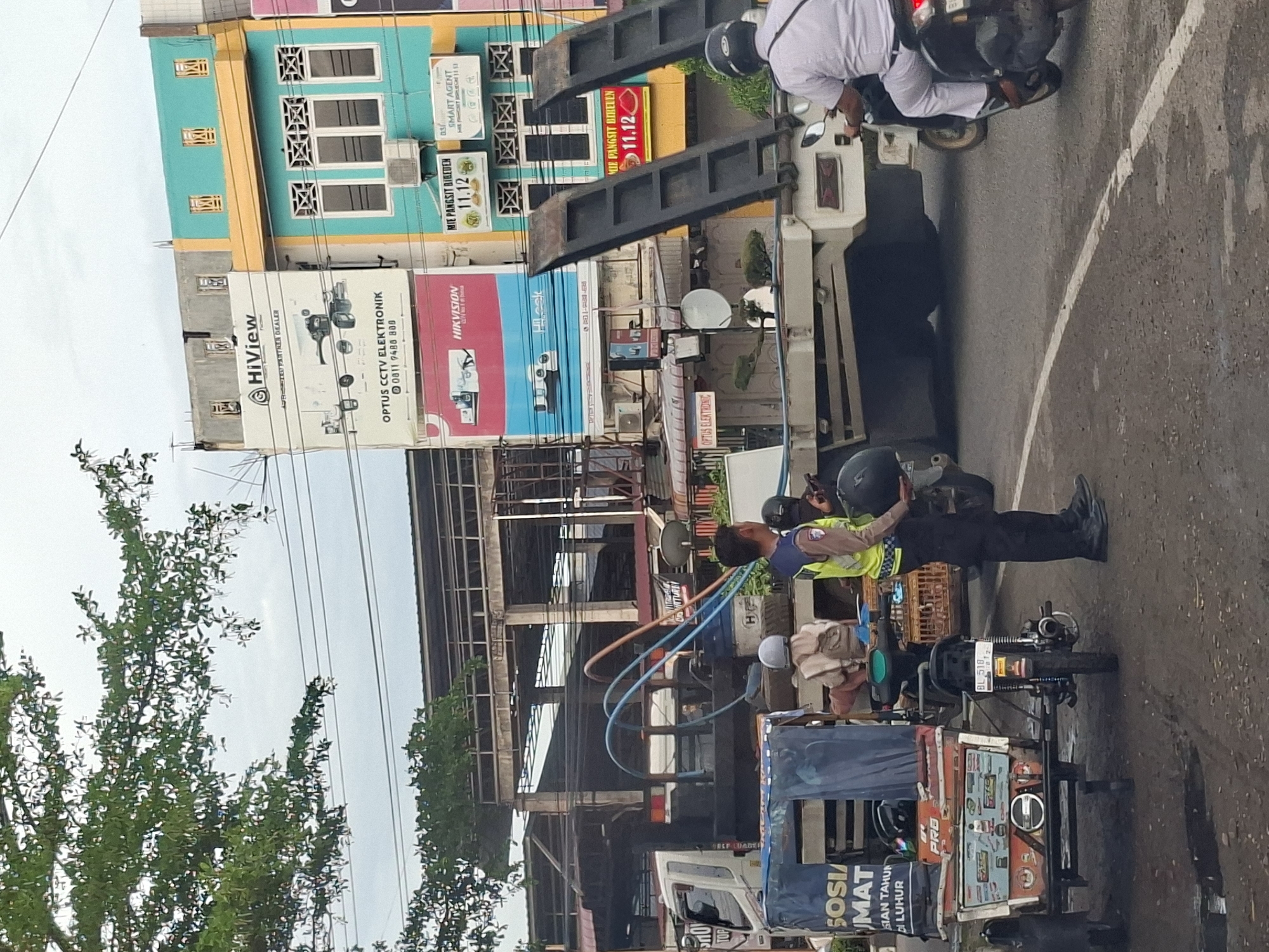 The image shows a street scene in Lhokseumawe, Indonesia, with a traffic incident or ongoing situation. In the foreground, a person in a yellow and blue vest, likely a traffic officer, stands holding a helmet. To the left, a three-wheeled vehicle with a blue cover is parked. Behind it, a large truck with two raised ramps is positioned across the road. A person wearing a light brown shirt and a helmet is seated on the truck. Another motorcycle is being ridden by a person in a white shirt and pants, heading away from the camera. The background features several multi-story buildings with colorful facades, displaying various advertisements including "HiView," "Optus CCTV Elektronik," "Hikvision," and "Mie Pangsit Bideden." The sky is overcast. The road surface is asphalt with some water patches and a white line marking.