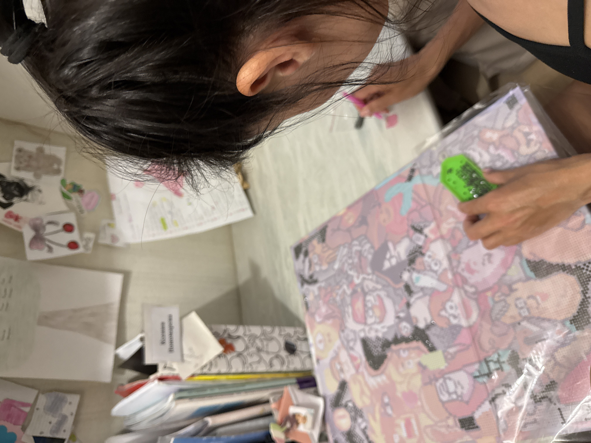 A woman with dark hair is hunched over a desk, focused on a diamond painting in a clear plastic wrapper. She is using a green applicator. The desk is cluttered with papers, drawings, a stack of books, and various other small items. A small paper on the desk is labeled "Koatee Romodjoglos." The location is Chaikovsky, Russia.