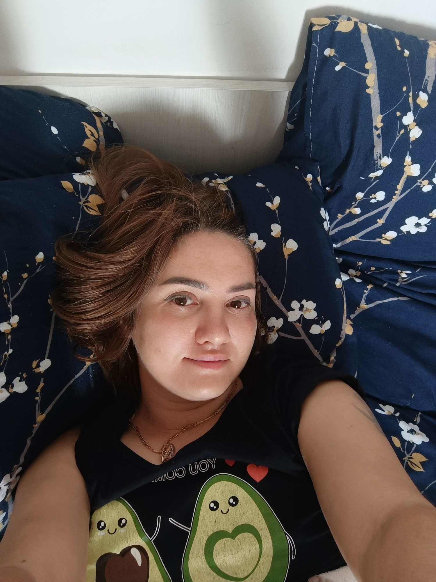 A woman with shoulder-length brown hair, wearing a black t-shirt with an avocado print, is lying on a bed with navy blue floral patterned sheets. She is looking at the camera with a slight smile. The scene appears to be an indoor setting, possibly a bedroom. There is no information about the location being in Taraz city administration, Kazakhstan.