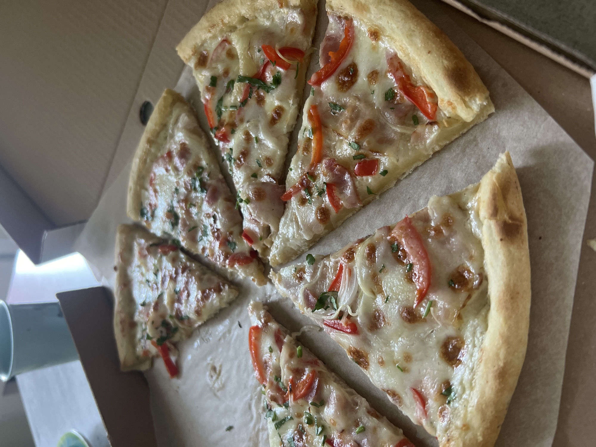The image shows a close-up, overhead view of several slices of pizza arranged in a cardboard pizza box. The pizza appears to have a white sauce base, cheese, red bell peppers, and what looks like ham or bacon, garnished with chopped green herbs. The crust is golden brown and slightly puffed. In the background, out of focus, is a light blue cup and part of a table, suggesting an indoor setting. There are no people visible in the image. The scene is lit by natural light, creating some shadows. The location is not specified in the image.