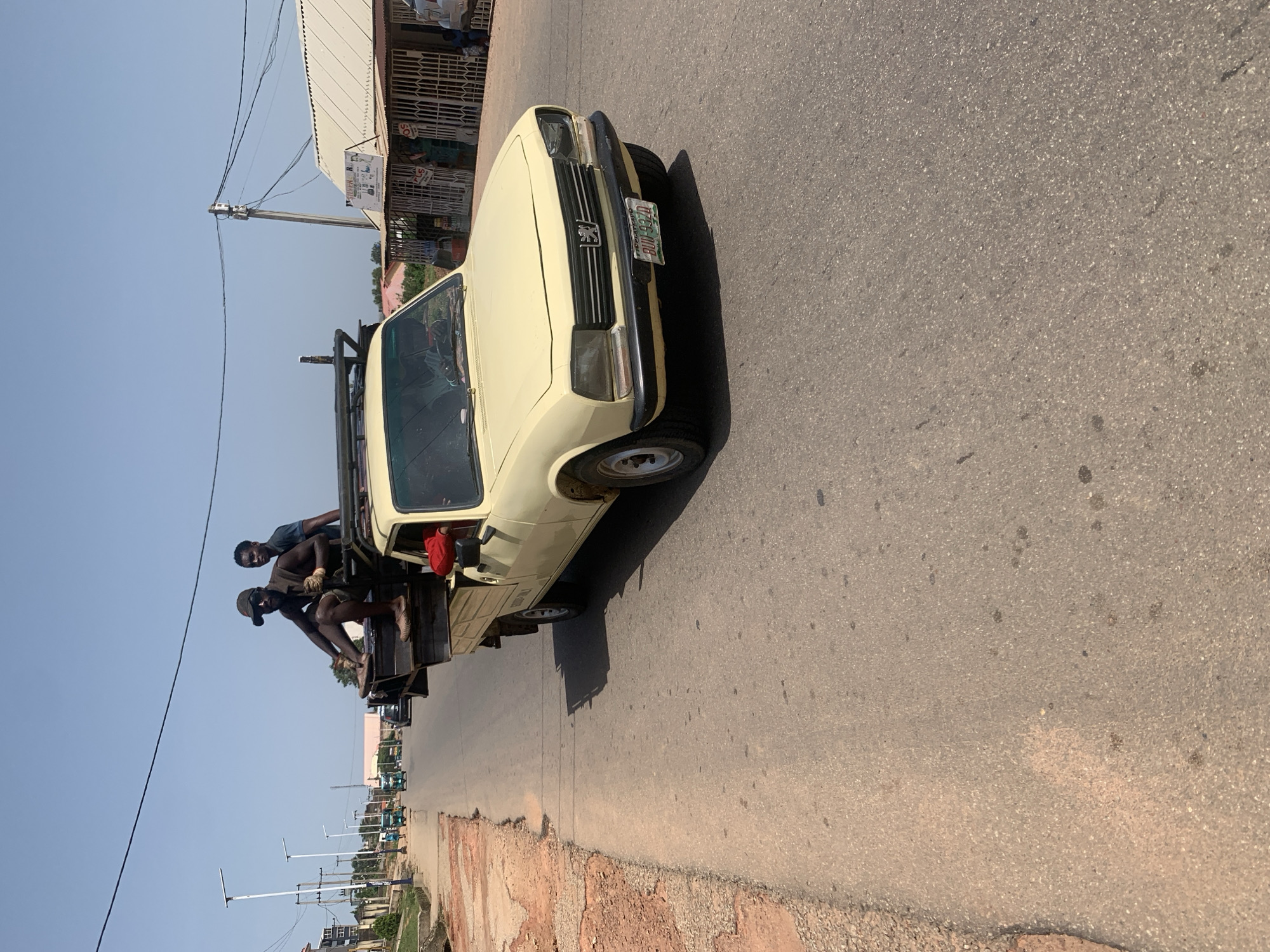 A light yellow Peugeot 504 pickup truck, modified with a flatbed for cargo, is parked on a paved road. The truck has a visible license plate that reads "ROD 997 D". Two men are seated on the flatbed of the truck, appearing to be resting or waiting. One man is wearing a dark t-shirt and light-colored shorts, with gloves on his hands. The other man is wearing a dark shirt and shorts and has a cap on. A portion of a person in a red shirt is visible in the driver's seat.
The truck is positioned on the right side of the road, which appears to be a two-lane asphalt surface. The road is lined with a disturbed dirt and gravel shoulder on the left side. In the background, a building with a corrugated metal roof and barred windows is visible, suggesting a commercial or market area. Utility poles and power lines run overhead. Further down the road, several other vehicles, possibly minibuses, are parked. The sky is clear and a bright blue, indicating a sunny day, likely in the late morning or early afternoon. The overall scene depicts a scene in Jos, Nigeria, with a focus on the truck and its occupants in a roadside setting.