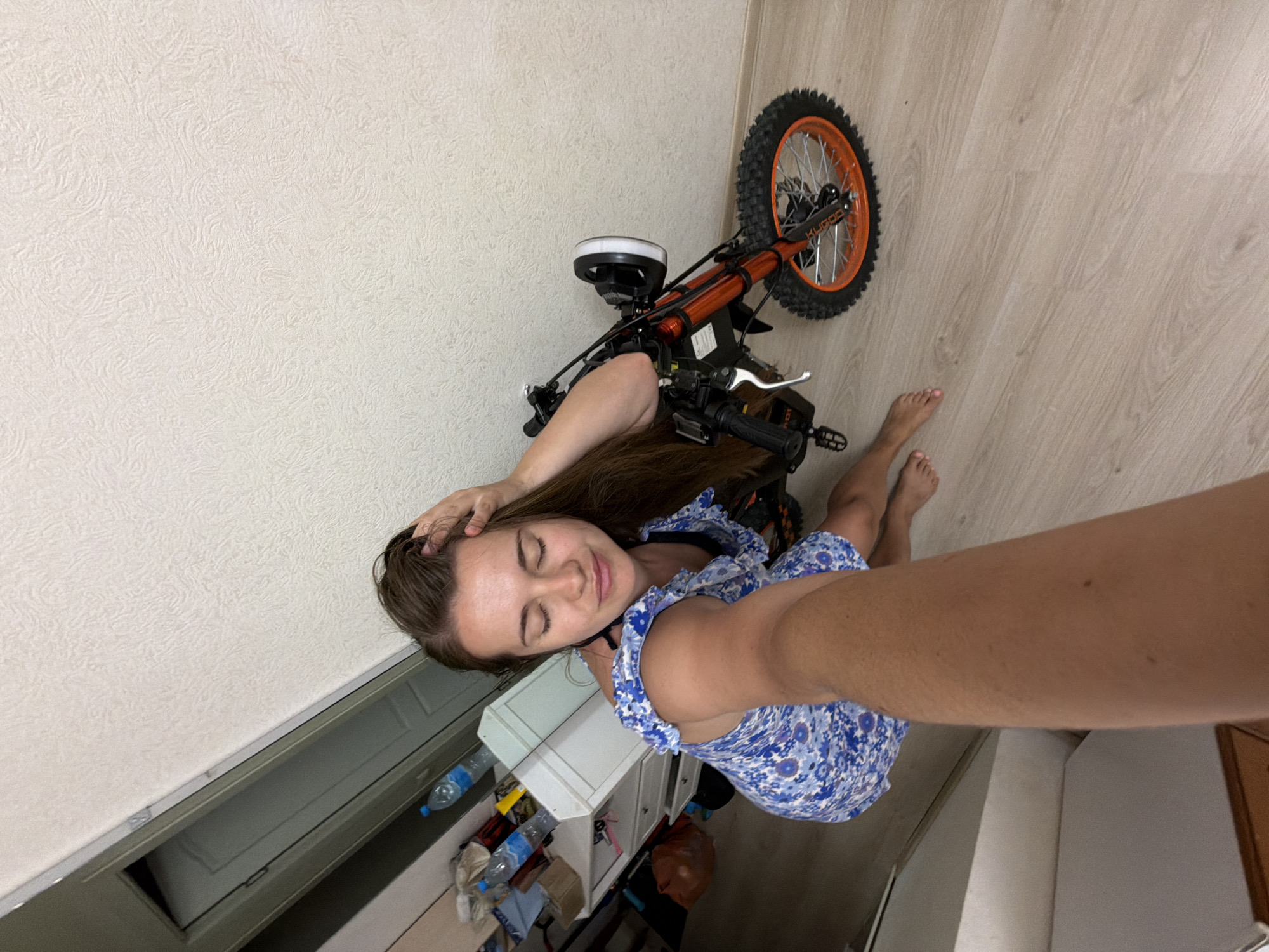 An elevated-angle indoor selfie captured in Sochi, Russia, depicts a female subject in her 20s or 30s. She is situated in the left-center of the frame, wearing a blue floral-patterned, off-the-shoulder dress or top, and is barefoot. Her eyes are closed, and a subtle smile is present on her face. Her left hand is raised to her head, touching her hair, while her right arm extends towards the camera, indicating she is holding the device. To the subject's right, a compact orange and black electric dirt bike or mini-bike is visible, featuring aggressive-tread tires, orange wheel rims, and a circular LED headlight with a white halo; "KUGOO" branding is discernible on the front suspension forks. The background on the left reveals a light-colored chest of drawers or cabinet, cluttered with several plastic water bottles, bags, and miscellaneous objects. Behind this, a larger white rectangular appliance, possibly a mini-fridge, is partially visible. The walls are textured and light-colored, and the floor is light-toned laminate wood. The scene presents a casual, domestic indoor environment.